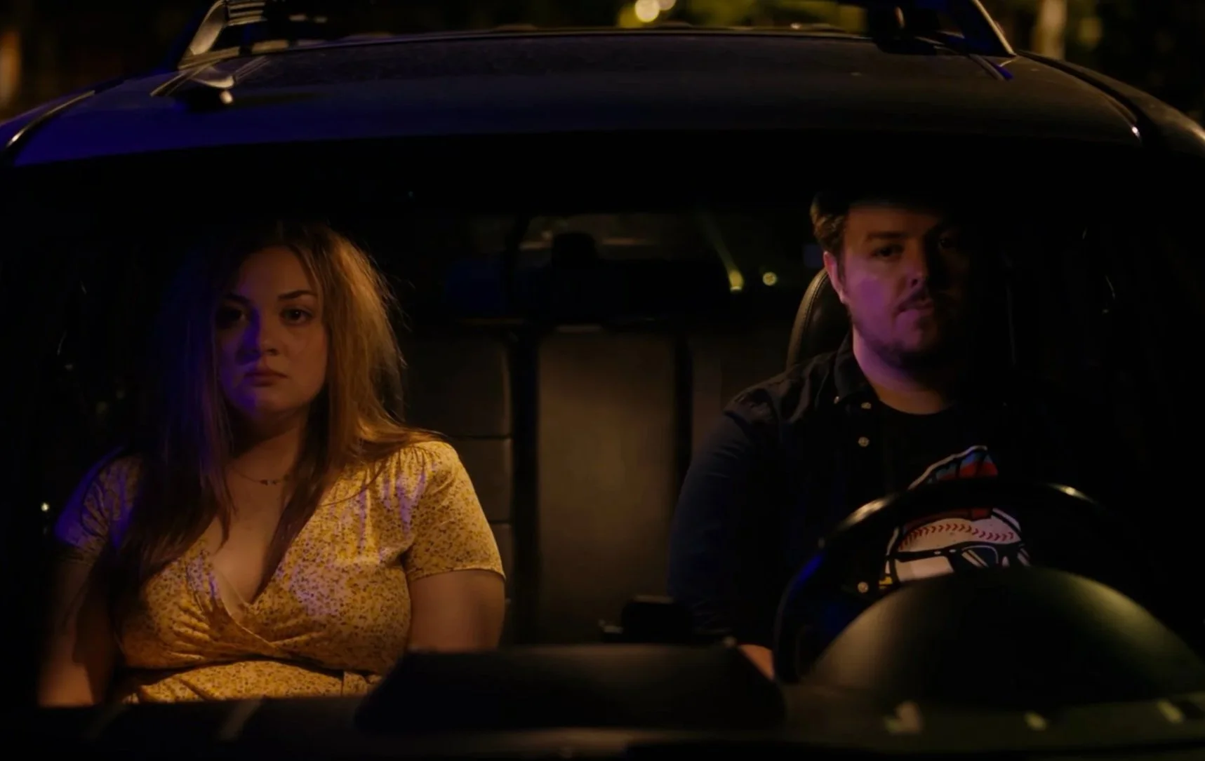 A young woman with long, messy hair in a yellow sundress and a young man with short hair and facial hair sitting in the front seats of a car at night. Lexi Putter & Jordan Black