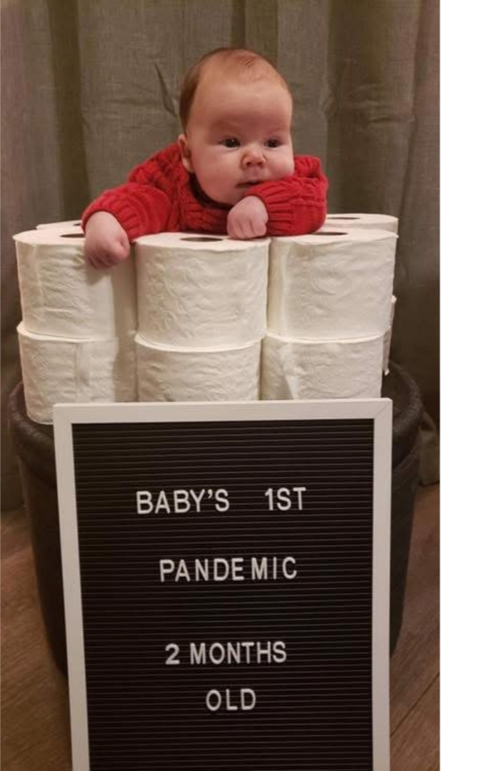 A baby in a red sweater lying on top of stacked toilet paper rolls with a sign in front that reads 'Baby's 1st Pandemic 2 Months Old'.