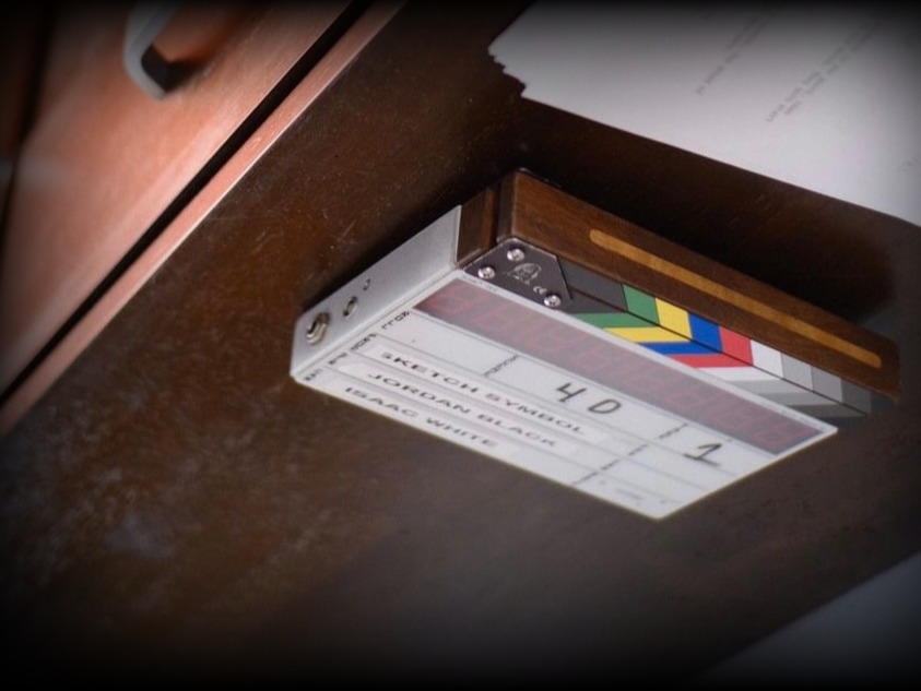 Clapperboard used in film production, for the show "Jordan Black: SKETCH Symbol" with color-coded lines on a wood surface.
