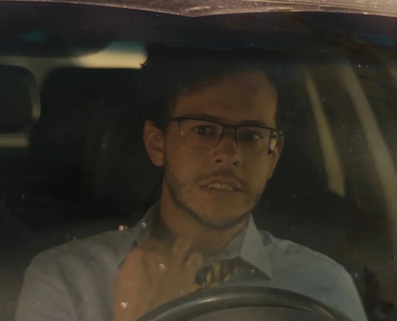 A man wearing glasses sitting in his car, seen through the windshield. He tugs at his shirt collar anxiously. Sam Campos