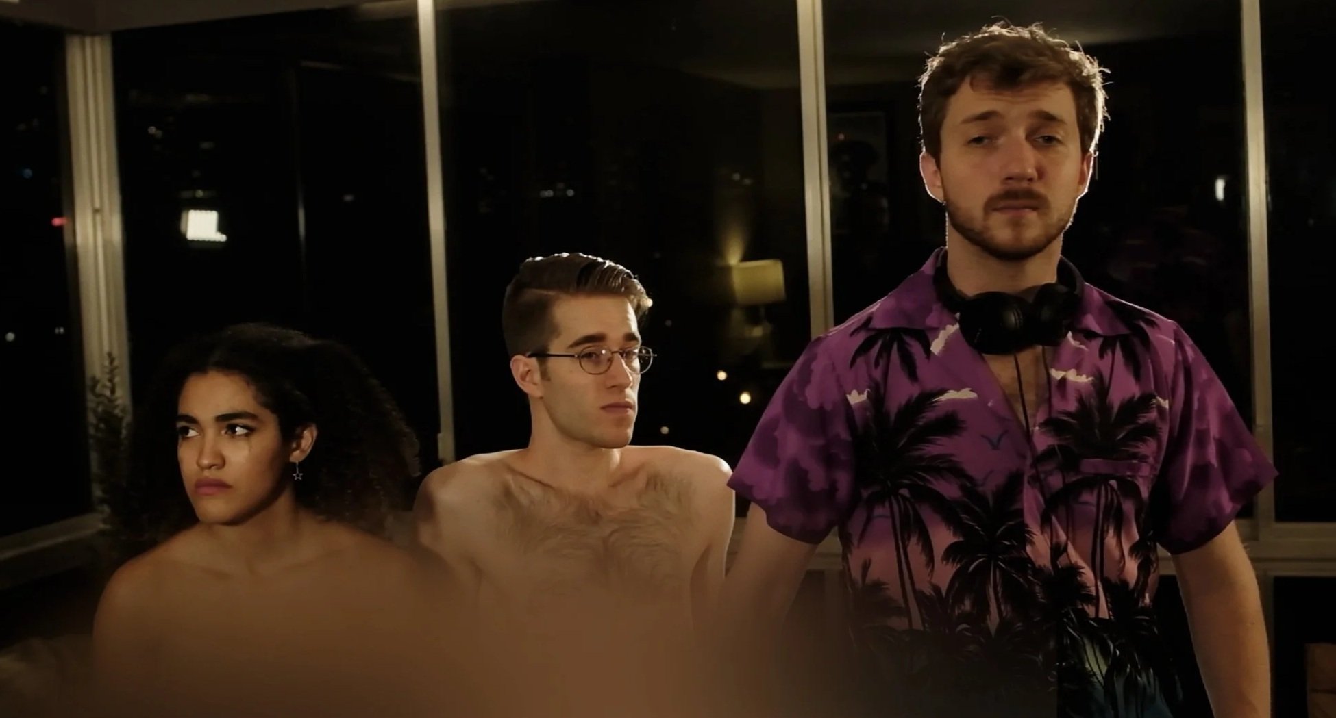 Three young adults at night, with a man in a purple Hawaiian shirt with black palm trees on the right, wearing headphones around his neck, and two blurry shirtless people on the left. Olivia Evans, Ben Southerland & Spencer Whipple