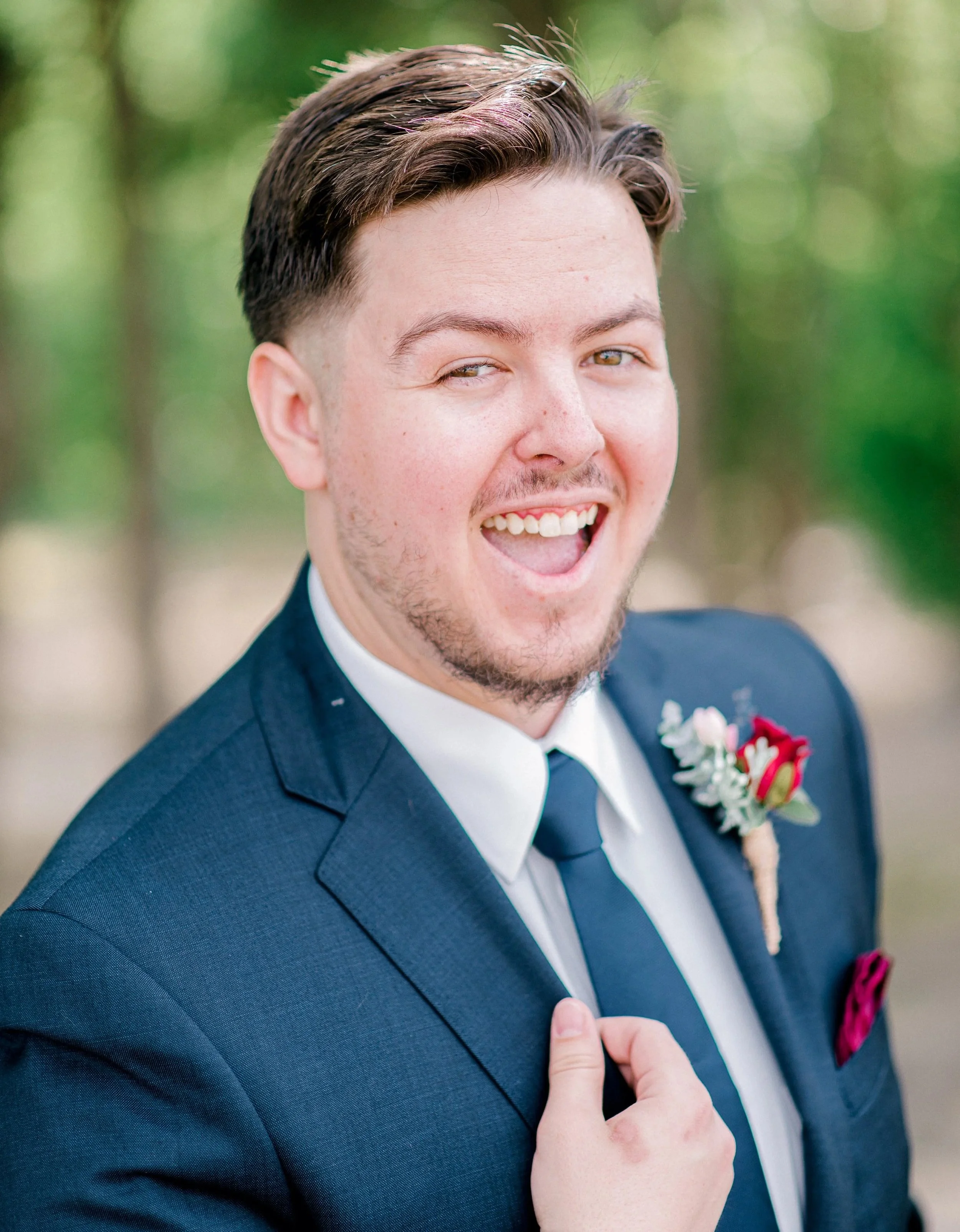 Close-up of a young man in a suit, smiling with his mouth open, outdoors with blurred green background.