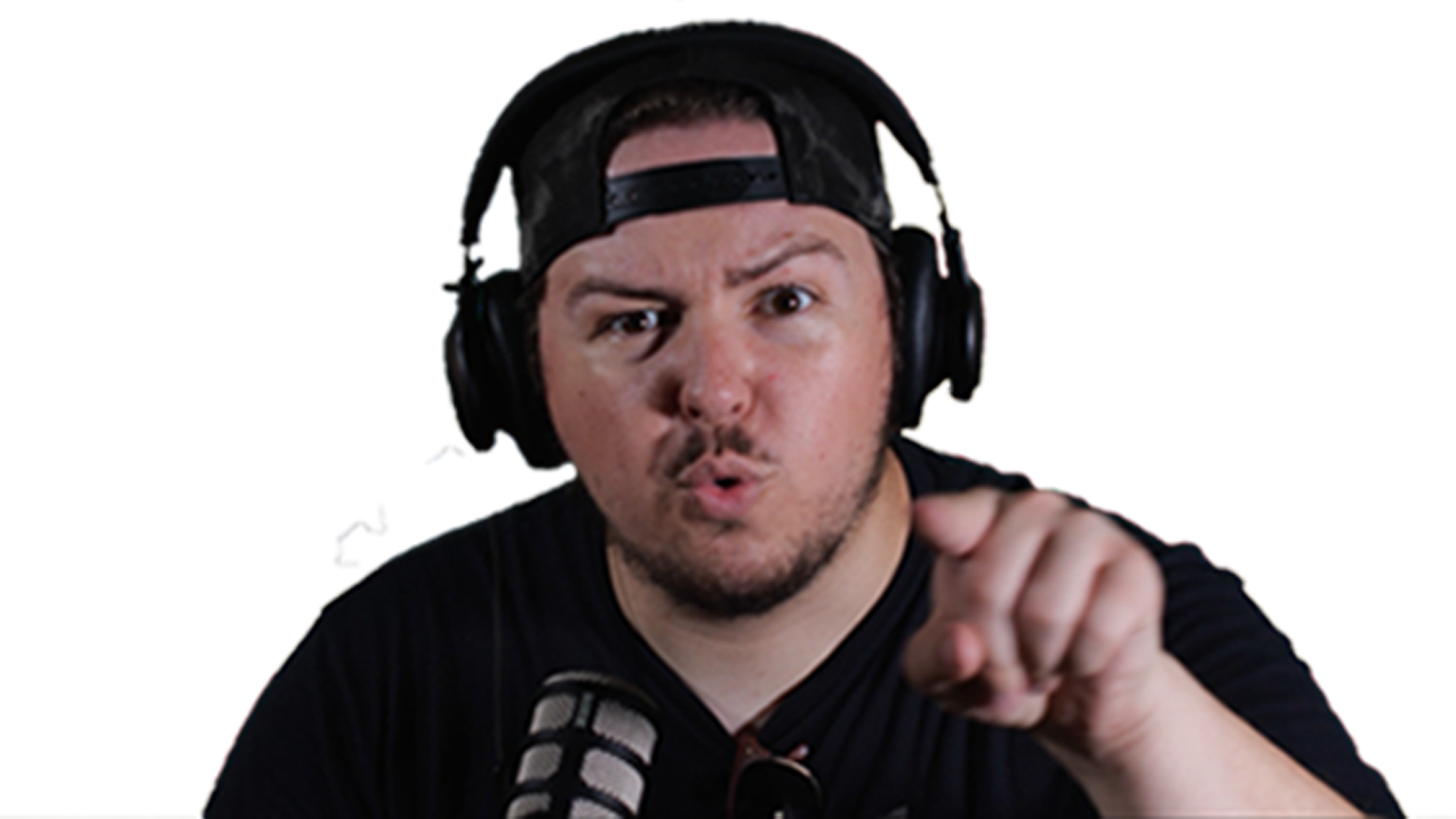 A young man wearing a black cap backwards and black headphones, looking intense, speaking into a microphone & pointing at the camera.