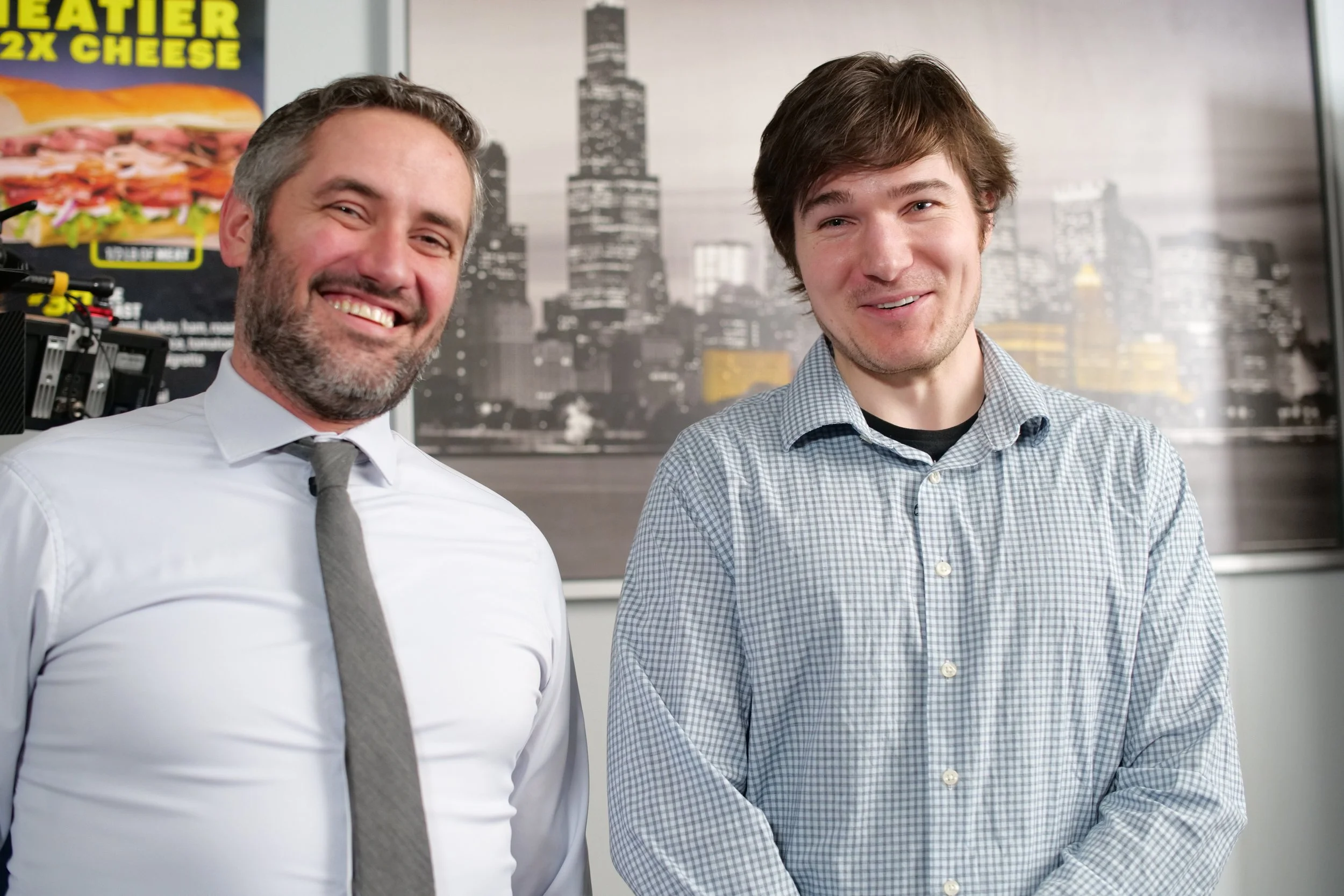 Two men, one with gray hair and beard wearing a white shirt and gray tie, and the other with brown hair wearing a checkered shirt, smiling and standing in front of a cityscape background with a tall building. Joe Fernandez & Steven Haas