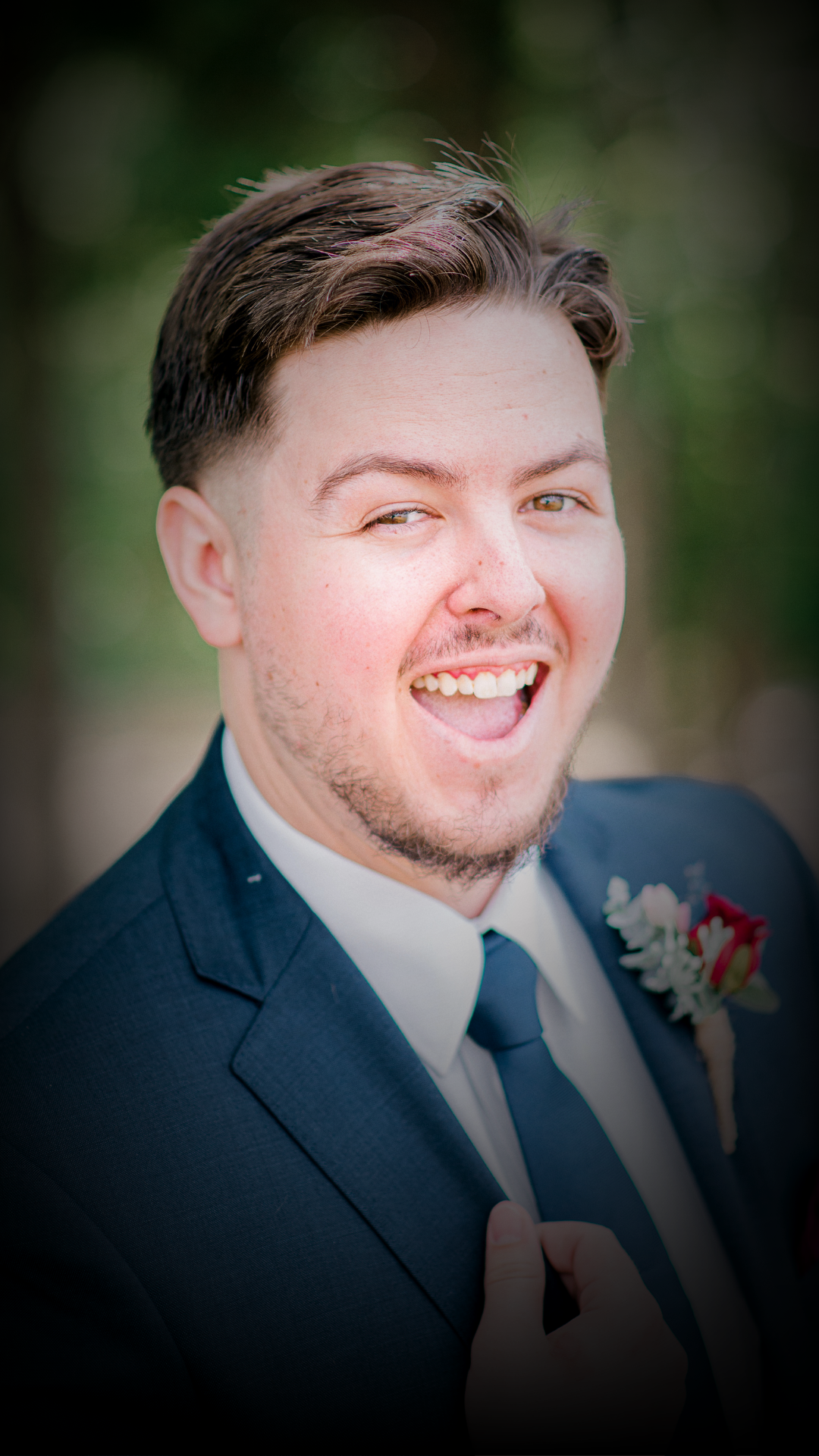 A young man in a blue suit smiling with an open mouth, outdoors with a blurred green background.