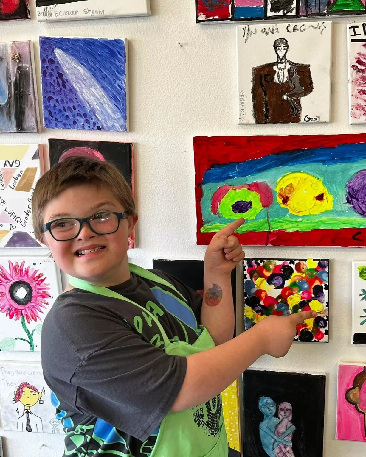 A young boy with glasses and a gray shirt smiling and pointing to a colorful abstract painting on a gallery wall, surrounded by other various children's artworks.