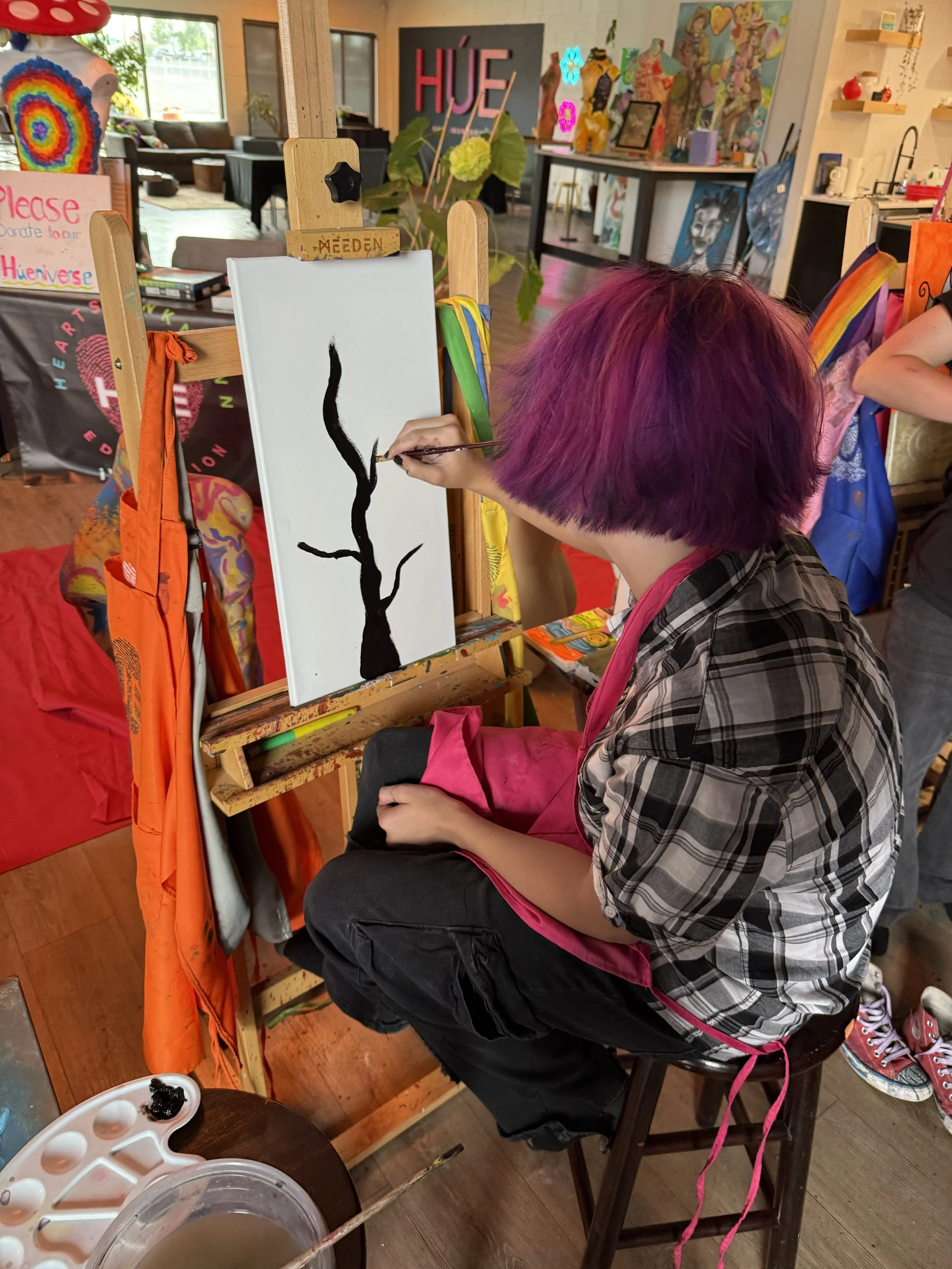 Person with purple hair painting a black tree branch on a white canvas in an art studio.