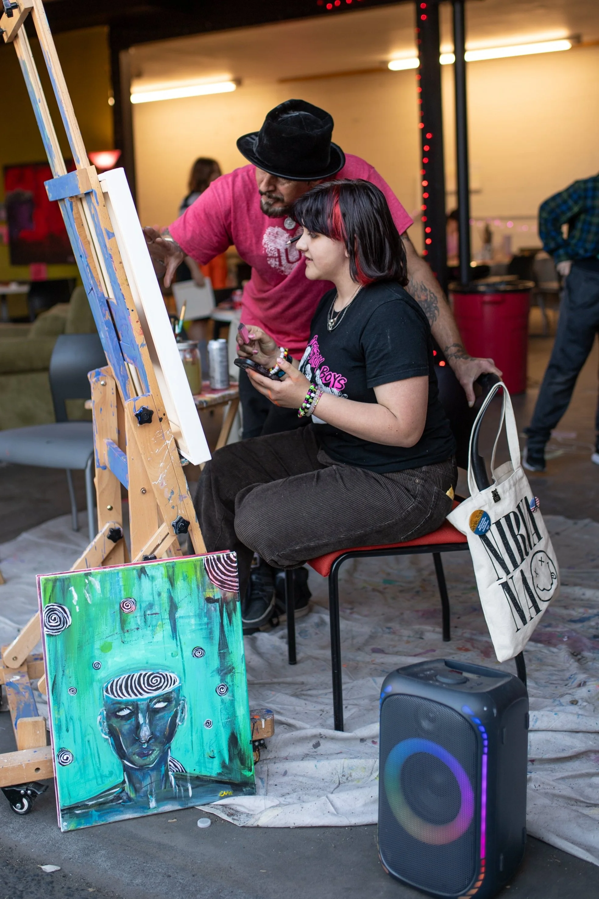 A woman with black and red hair sitting on a chair, using a smartphone, while an artist with a beard and hat looks at her. A painting of a woman with swirling patterns in green and blue tones is on the floor in front of them. There is a speaker on the floor near the woman, a canvas on an easel, and other people and art supplies visible in the background.