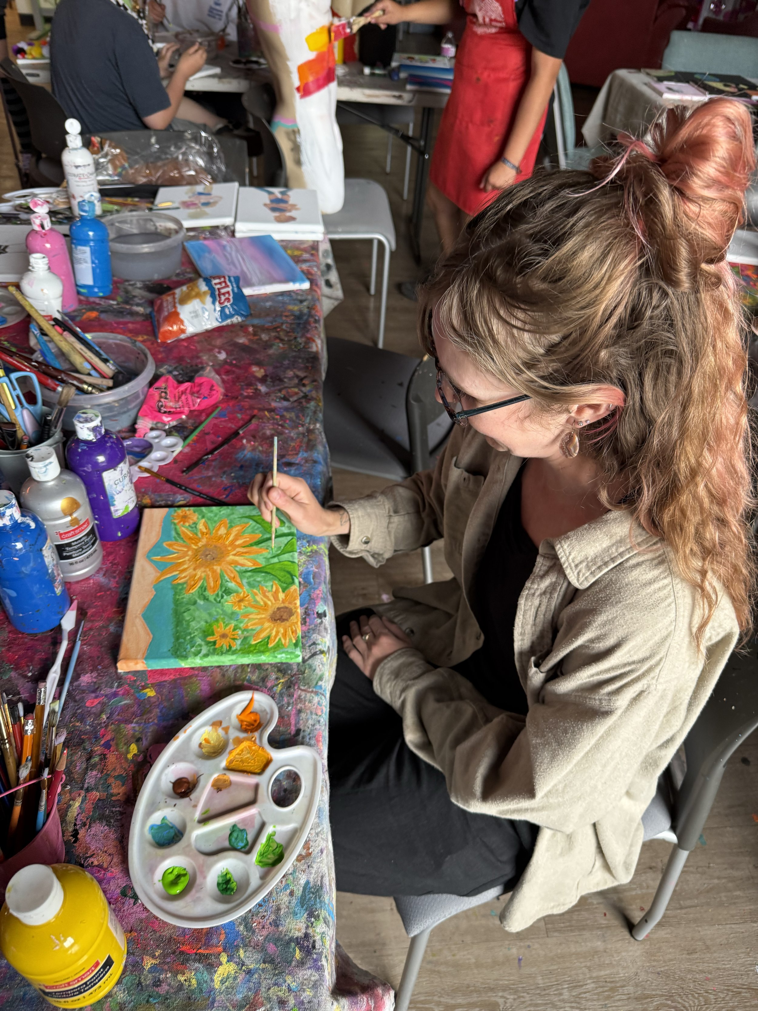 A woman with curly hair and glasses sitting at a table, painting a sunflower on a canvas with bright yellow and orange colors. The table is covered with art supplies including paints, brushes, and a palette with various colors. In the background, there are other people working on art projects in a studio or classroom setting.