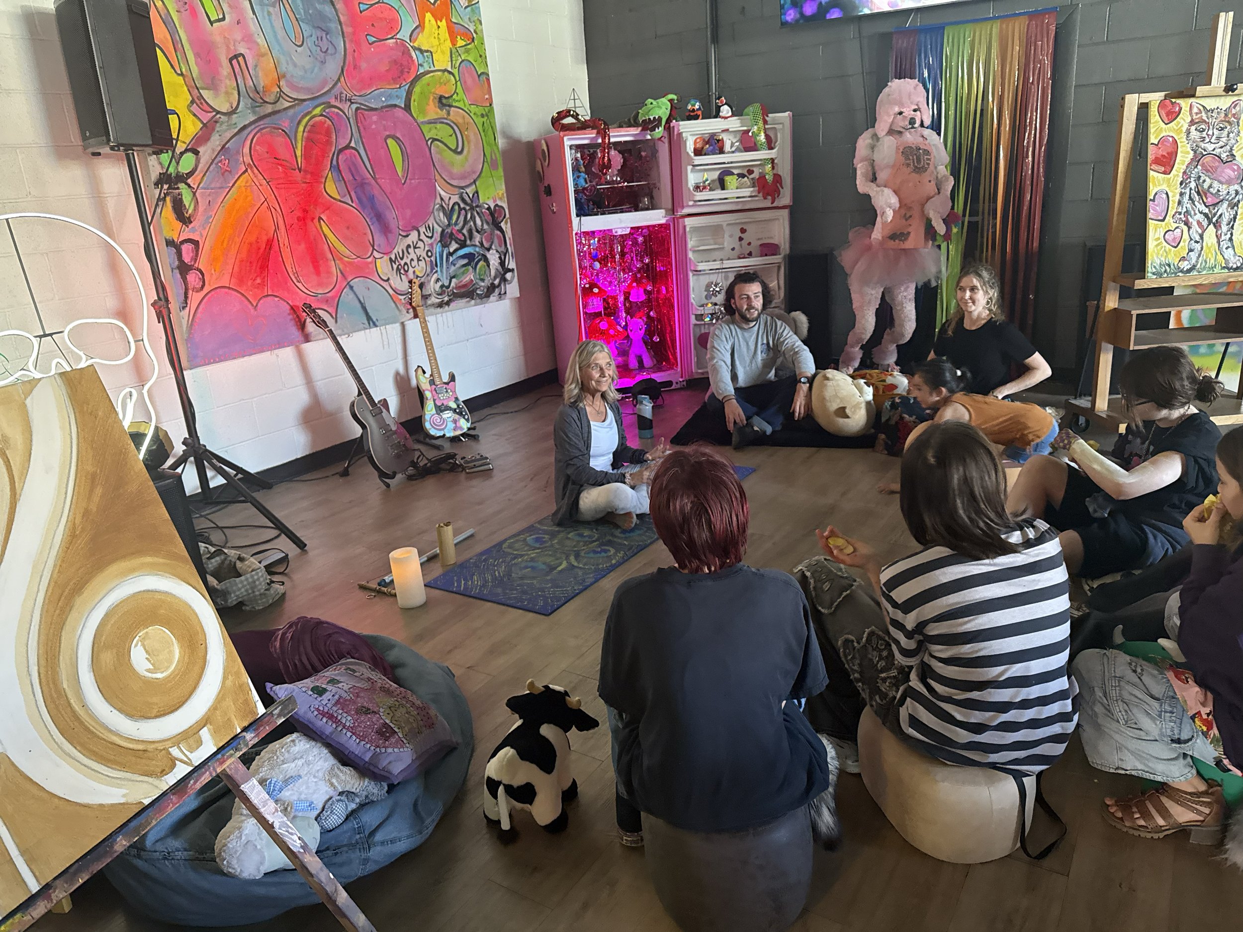 A group of people sitting on the floor in a colorful art studio or classroom, surrounded by paintings, guitars, stuffed animals, and art supplies. There is a large pink stuffed animal, artwork on the walls, and a small girl standing among the seated individuals.