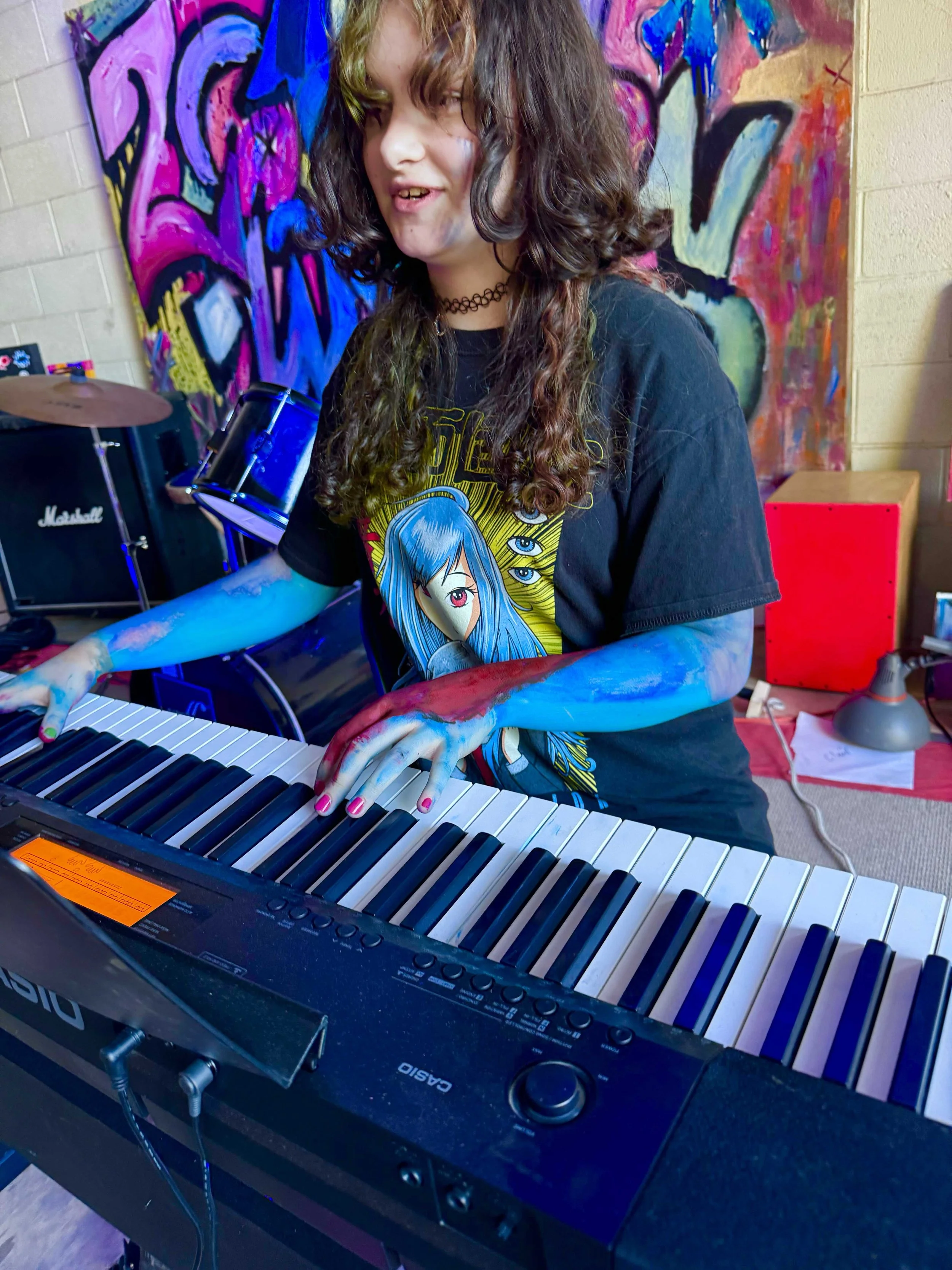A person with long, curly hair playing a digital keyboard in a room decorated with vibrant graffiti art on the wall behind them. The person has a choker necklace and is wearing a black T-shirt with colorful animated characters on it.