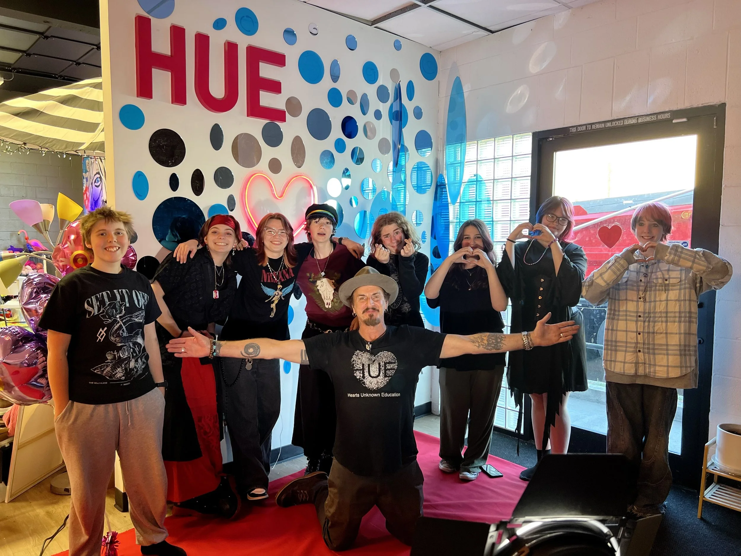 A group of people posing for a photo indoors in front of a colorful wall decorated with the word 'HUE,' a heart, and various sized circles. They are smiling and making gestures, some with their hands on each other's shoulders or forming heart shapes with their fingers. There are balloons and a band at their feet, and bright natural light coming through a window.