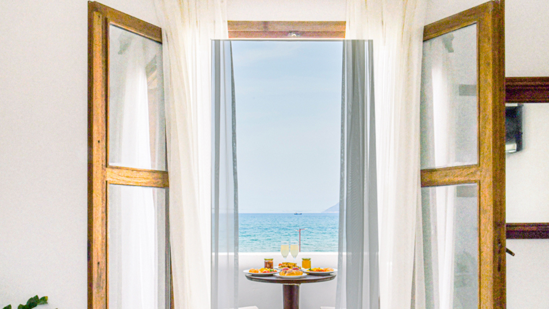 Open window with white curtains overlooking the ocean, with a small table outside holding breakfast, including two glasses of champagne, plates of food, and jars of jam.
