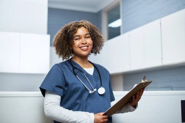 A young female nurse with curly hair wearing blue scrubs and a stethoscope holding a clipboard and smiling.