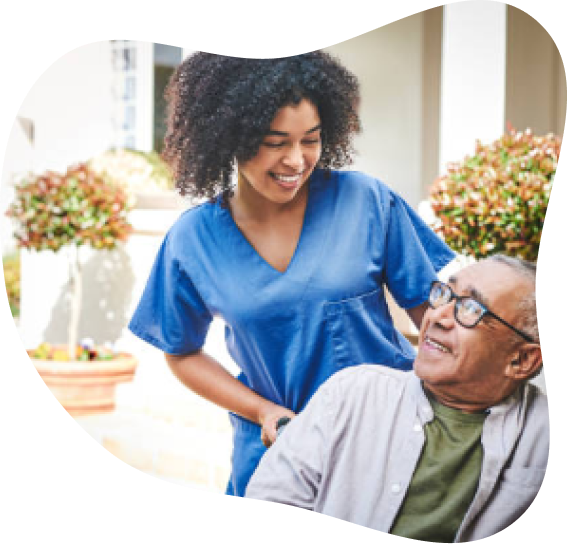 A nurse or caregiver talking to an elderly man in a bright room with potted plants in the background.