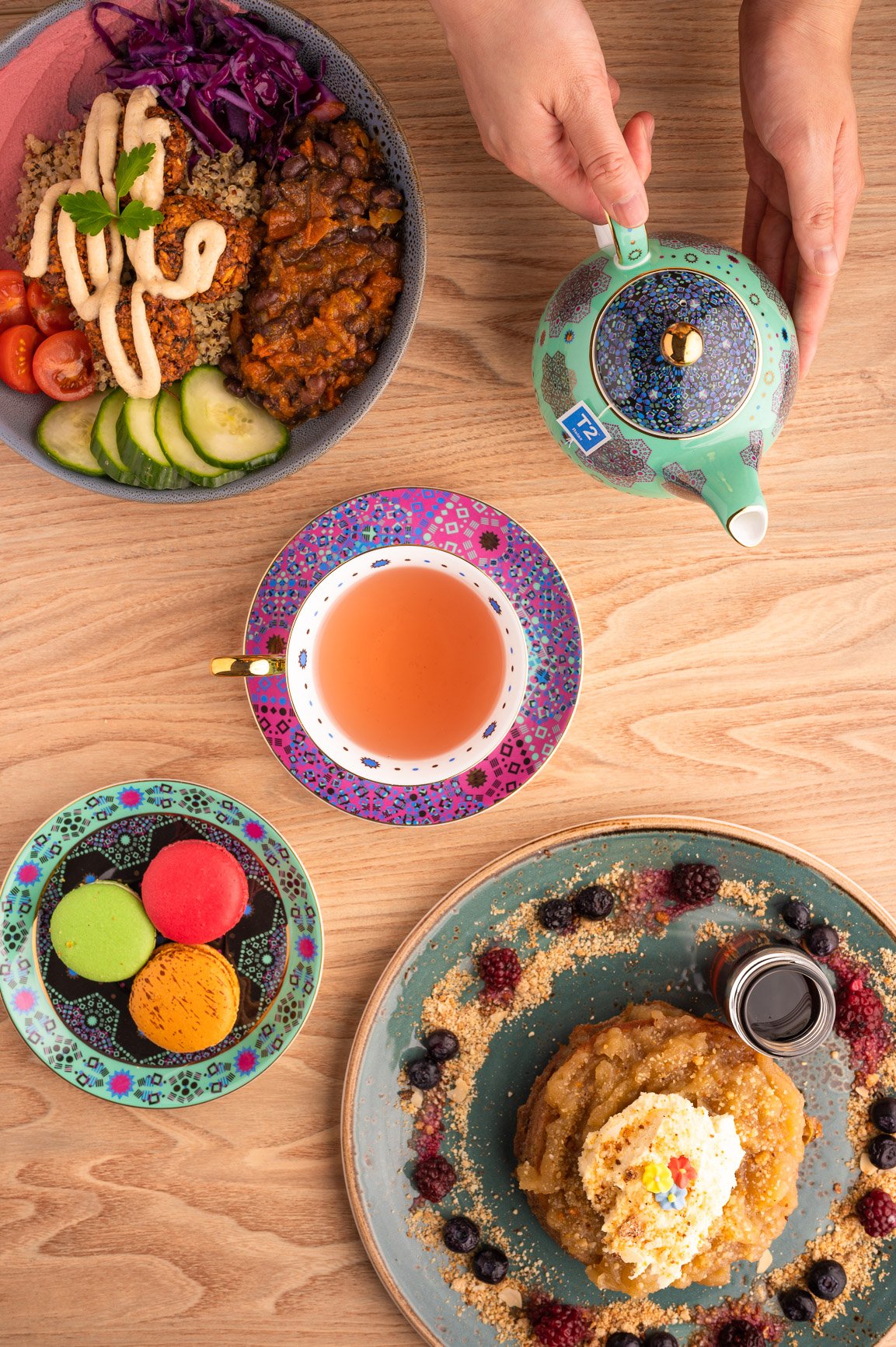 Overhead view of a wooden table with a bowl of quinoa, vegetables, and sauce, a colorful cup of tea, a plate with three macarons, and a dish of dessert topped with cream and berries, accompanied by a teapot.