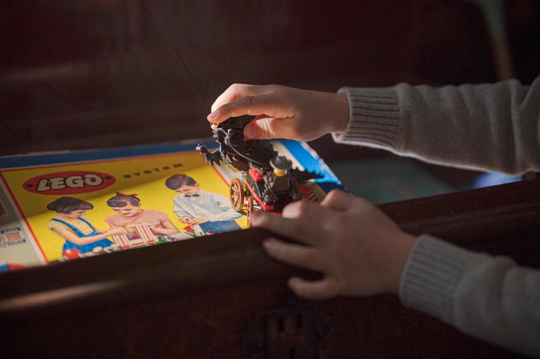 Child holding a LEGO carriage over a vintage LEGO set box featuring three children playing, with a colorful yellow background.