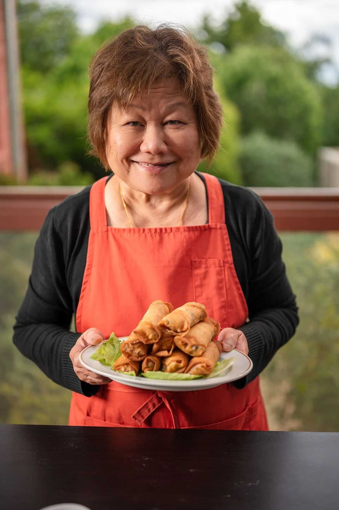 A smiling person wearing a red apron holding a plate of spring rolls.