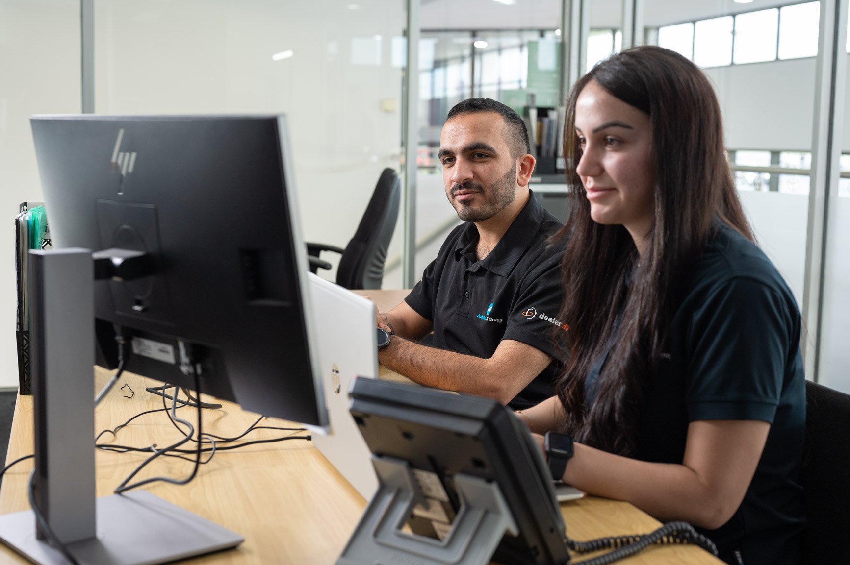 Two people working at a desk with computer monitors in an office.
