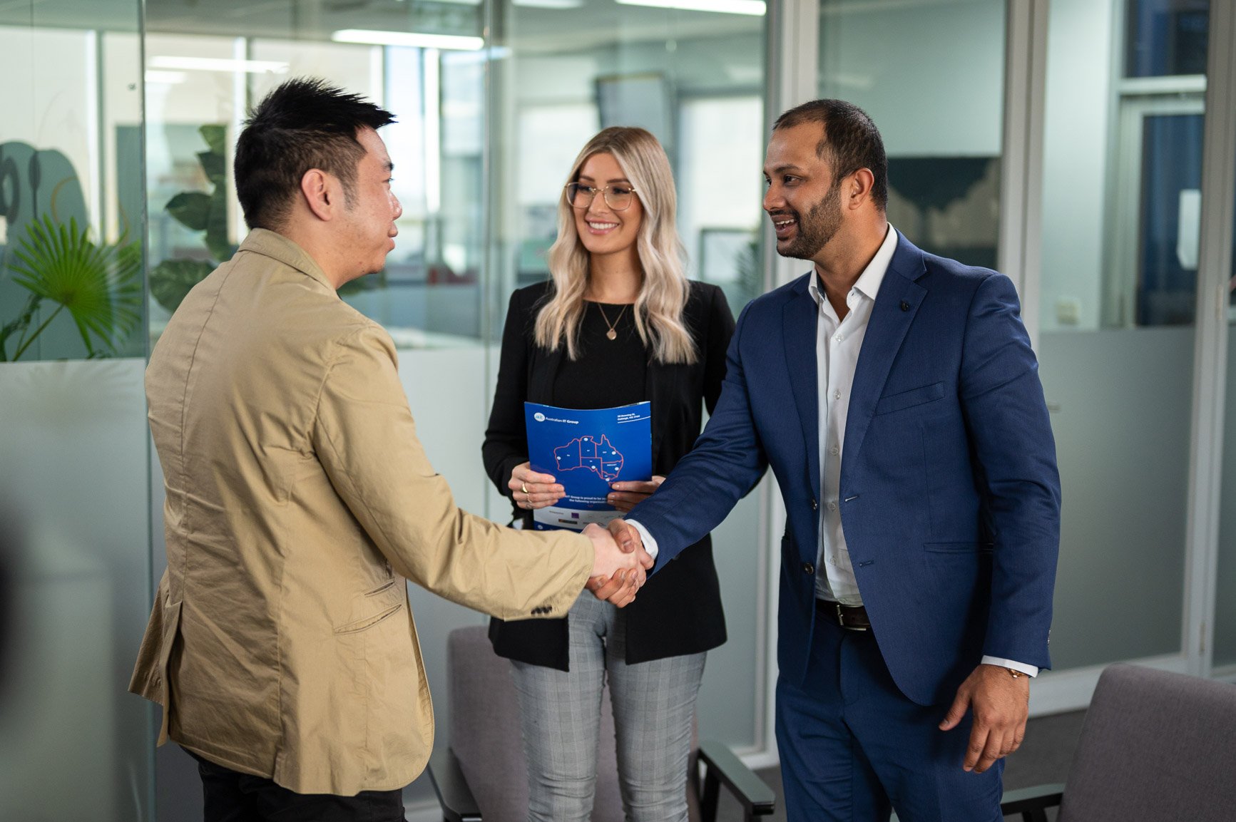 Three professionals in an office, two men shaking hands, a woman holding a brochure.