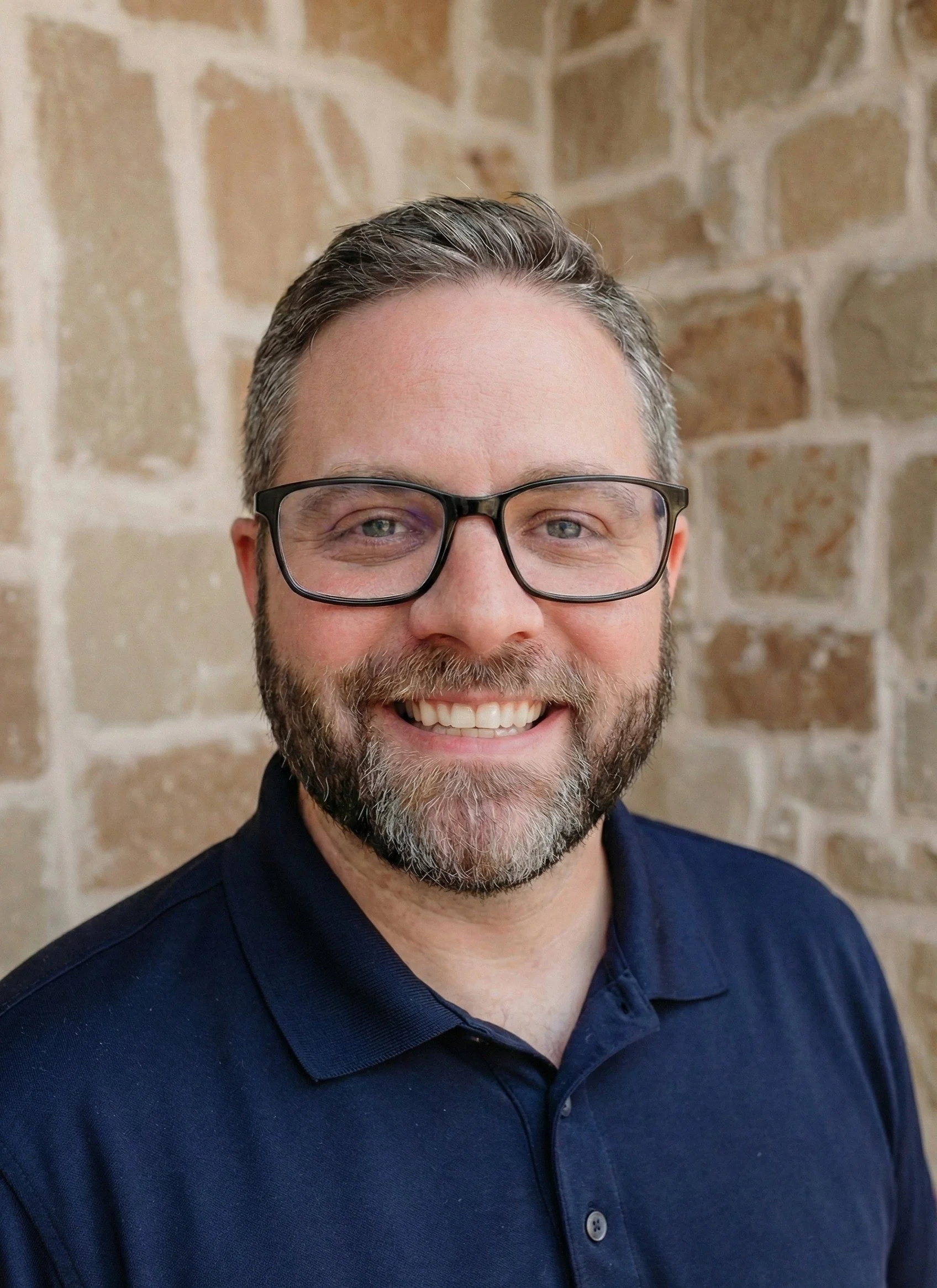 A smiling man with glasses, a beard, and short hair, wearing a navy polo shirt, standing in front of a tan-colored brick wall.