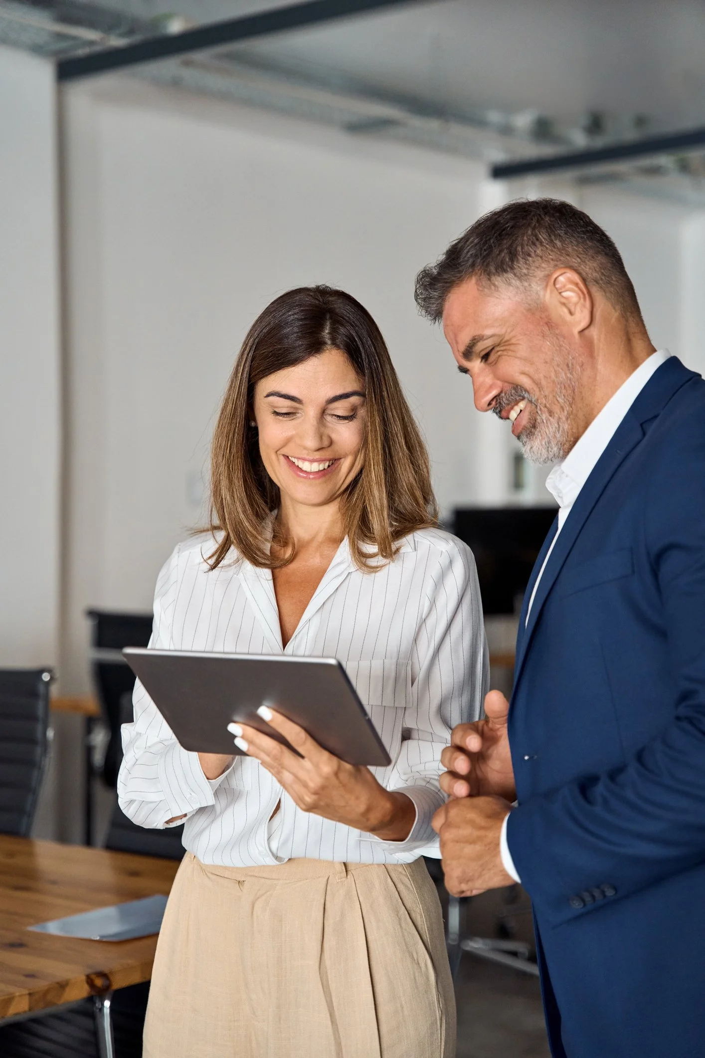 A man and a woman in business attire are standing together in a modern office, looking at a tablet device and smiling.
