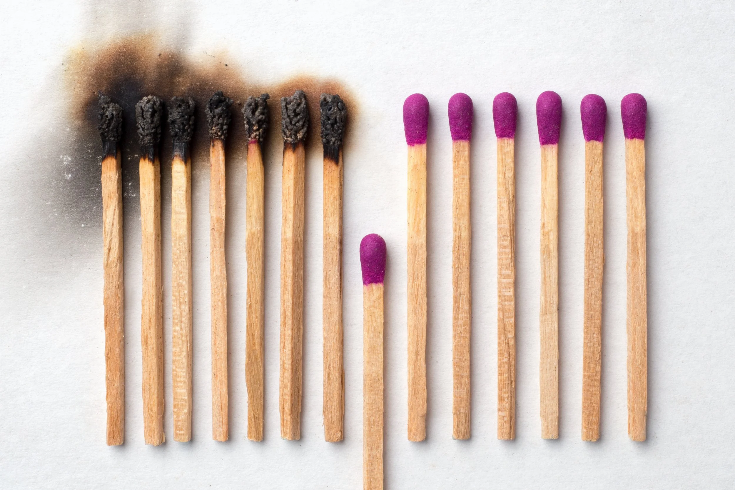 Six matches on the left are burnt and blackened, with burned heads, while seven matches on the right are unburned with purple heads, all aligned on a white surface.