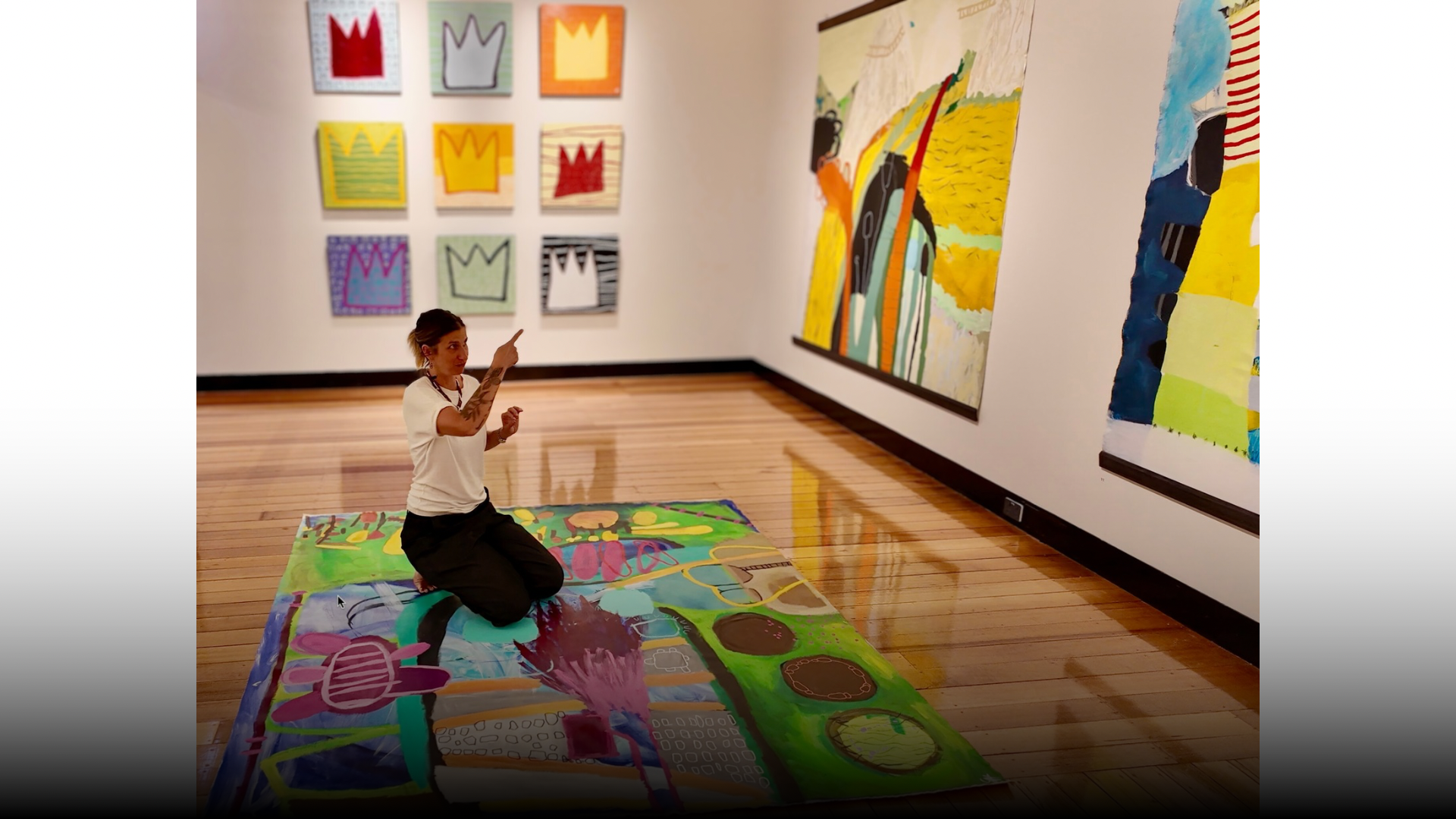A woman kneeling on a colorful painted floor in an art gallery, pointing at paintings on the wall. The gallery has a wooden floor and white walls, with various abstract and colorful artworks displayed.