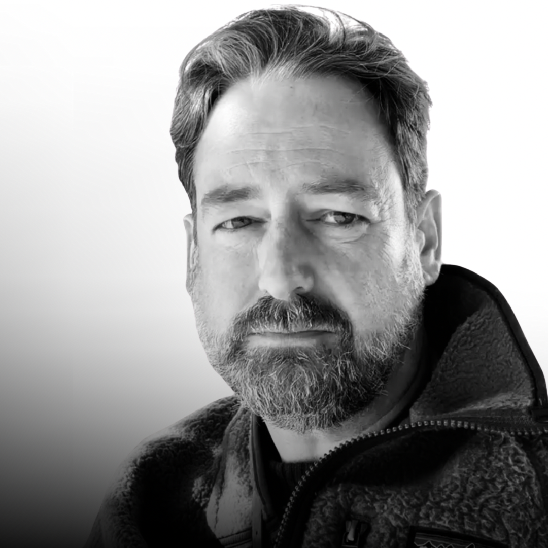 Black and white close-up of a middle-aged man with a beard and mustache, wearing a zippered jacket with a raised collar, looking at the camera with a serious expression.