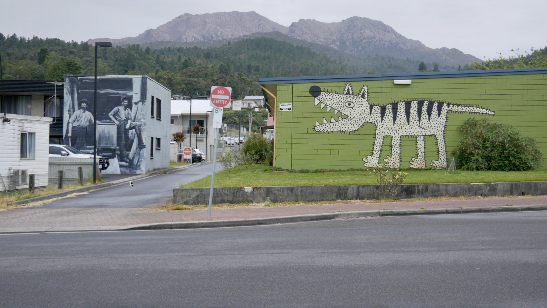 Street view featuring a green building with a large, cartoonish dinosaur mural, a black and white mural of two men on a neighboring building, and surrounding houses with mountains in the background.