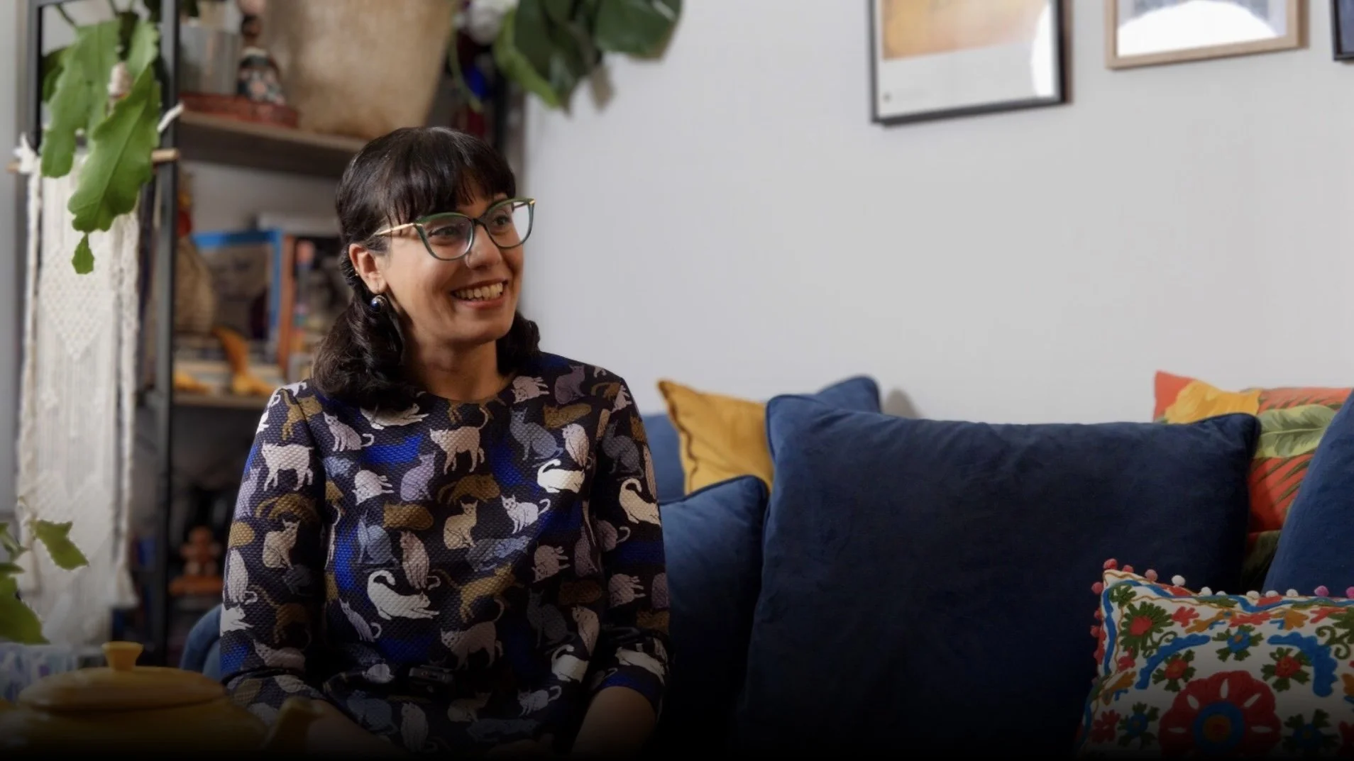 A woman with dark hair, glasses, and earrings smiling while sitting on a couch in a living room with colorful cushions and artwork on the wall.