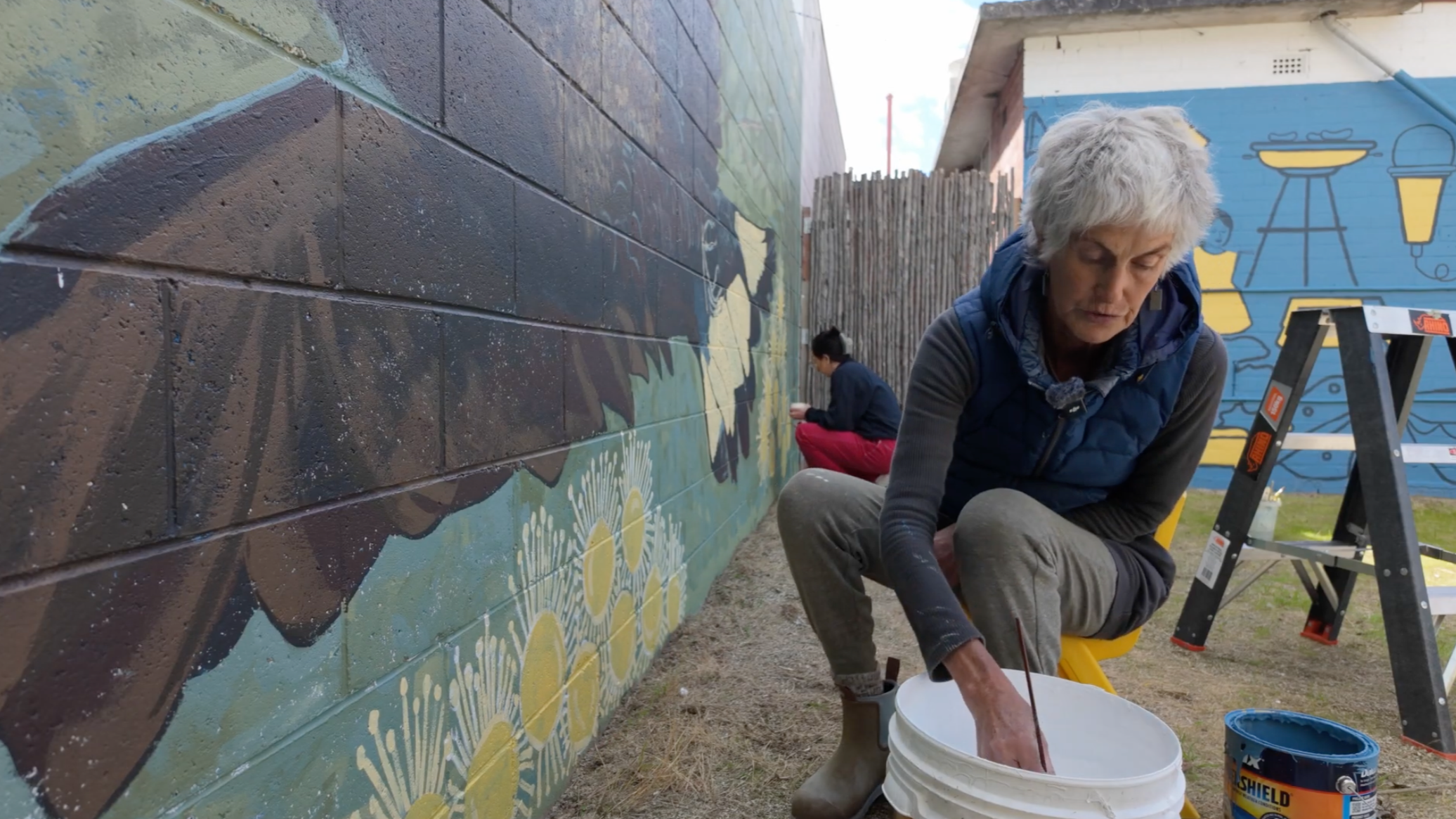 Two women painting a mural on a brick wall outdoors, one in the foreground with gray hair and a blue vest, and another in the background sitting on the ground. There are paint buckets and a ladder nearby.