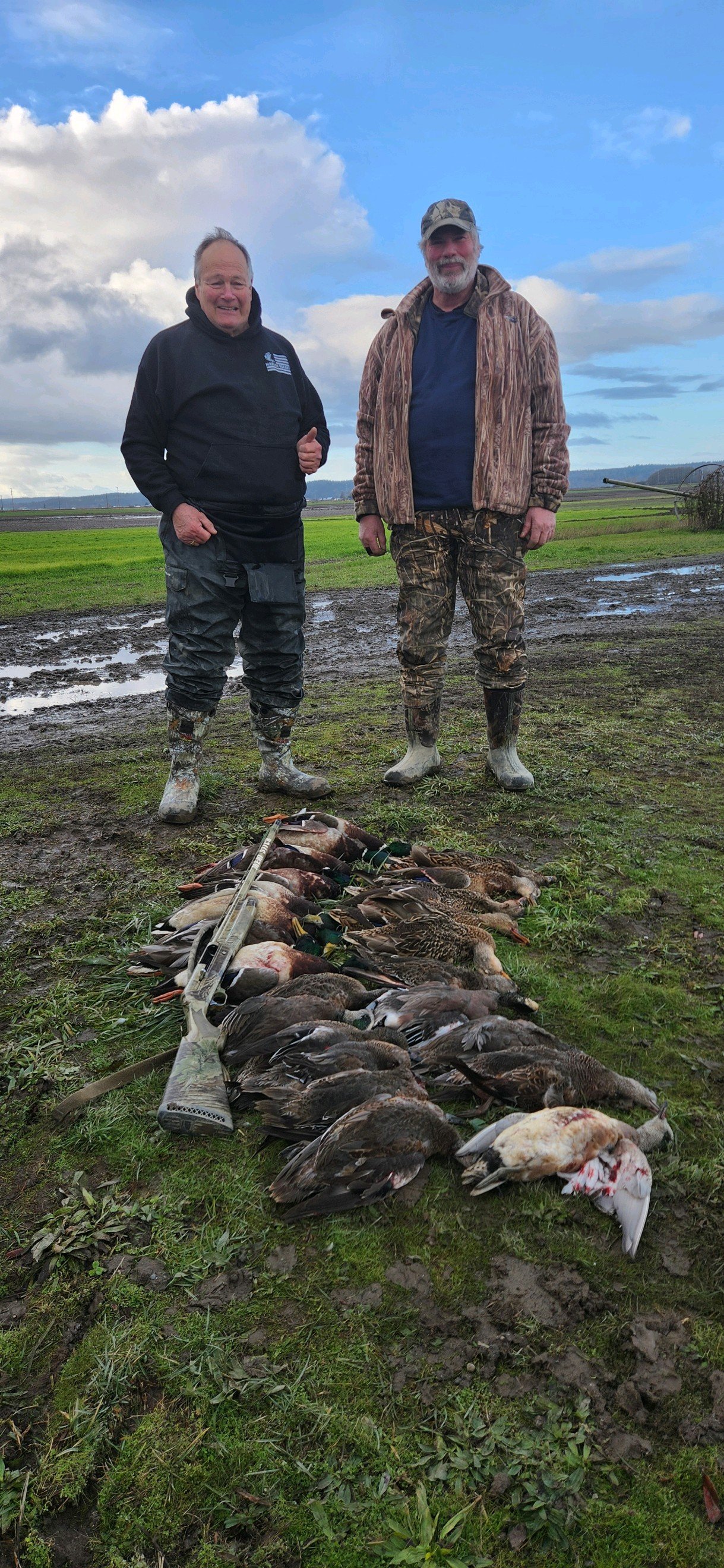 Two men standing outdoors on a muddy field, smiling at the camera. They are dressed in camouflage and outdoor clothing. In front of them, a lineup of ducks and other waterfowl is laid on the ground, with a rifle resting among the birds. The backgroun