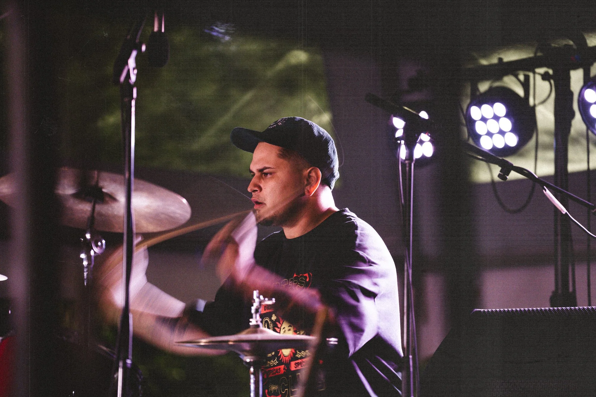 A young male drummer wearing a black cap, black shirt, playing drums on stage with purple stage lights.