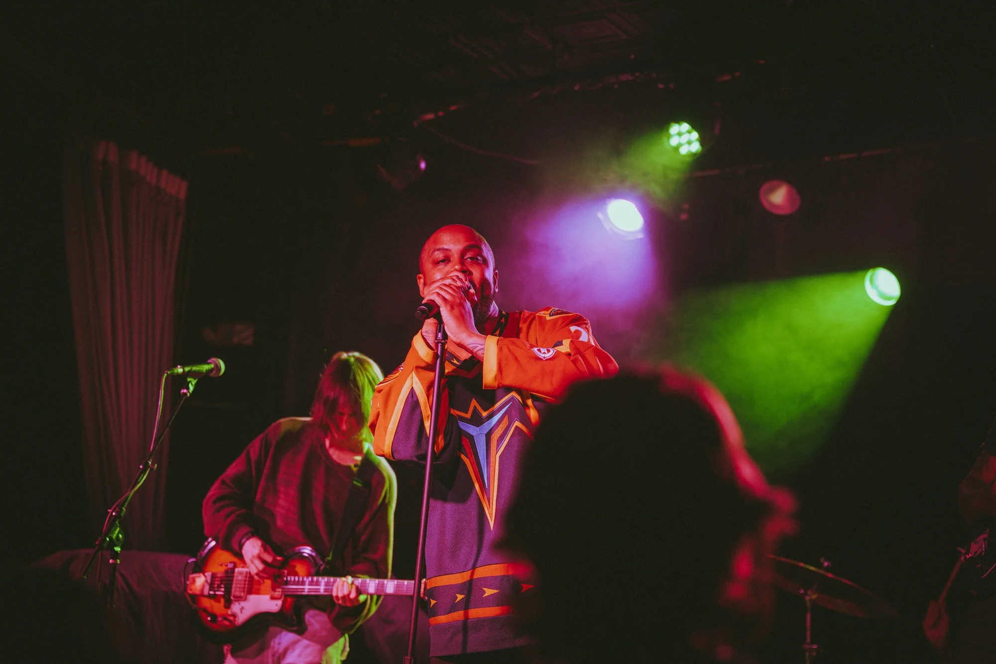 Male singer in colorful sports jersey singing into microphone on stage, with guitarist playing nearby, under colorful stage lights.