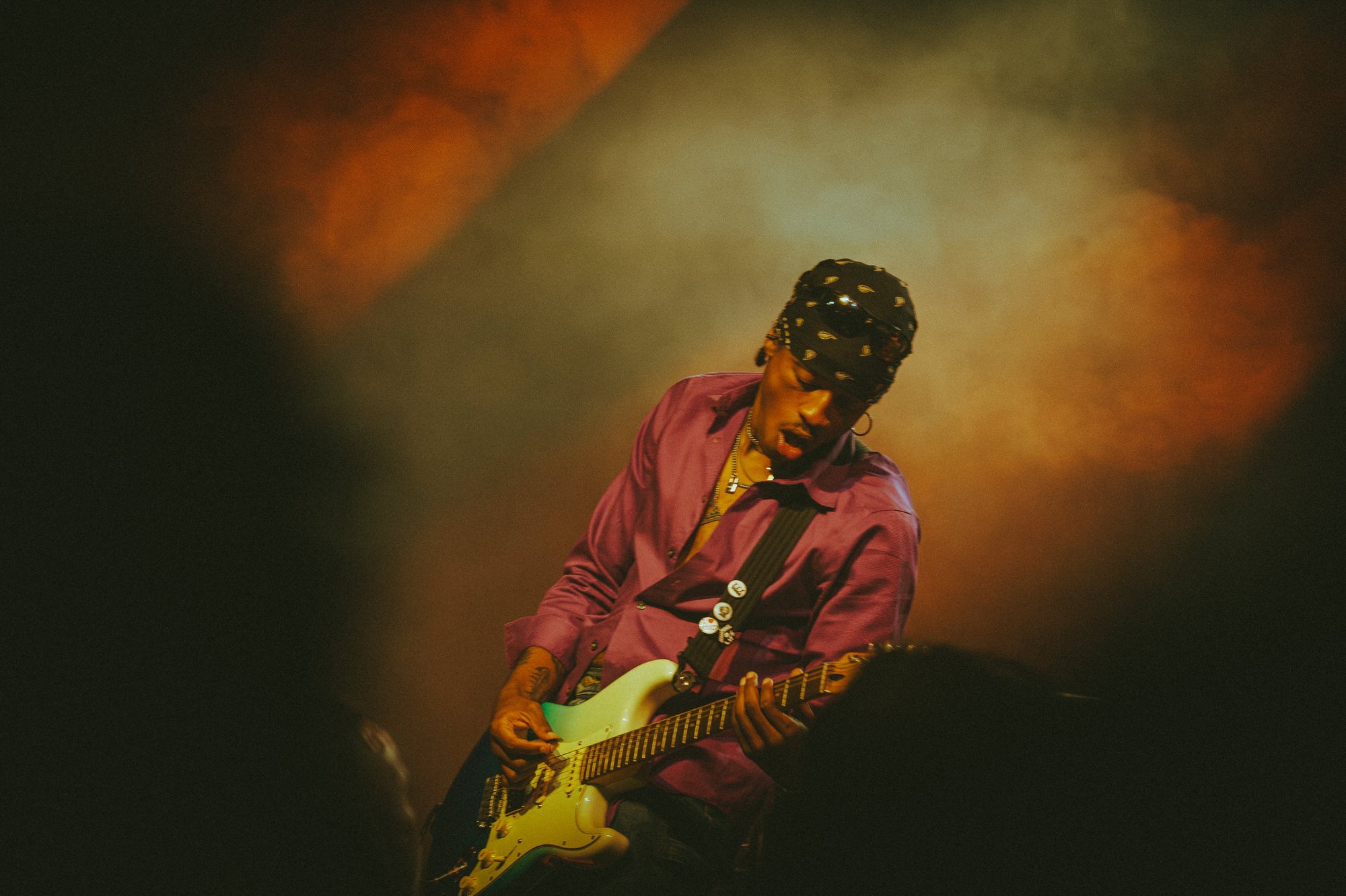 A person wearing a black bandana with white patterns, a pink shirt, and jewelry, playing an electric guitar in a dimly lit stage environment with warm colored lighting.