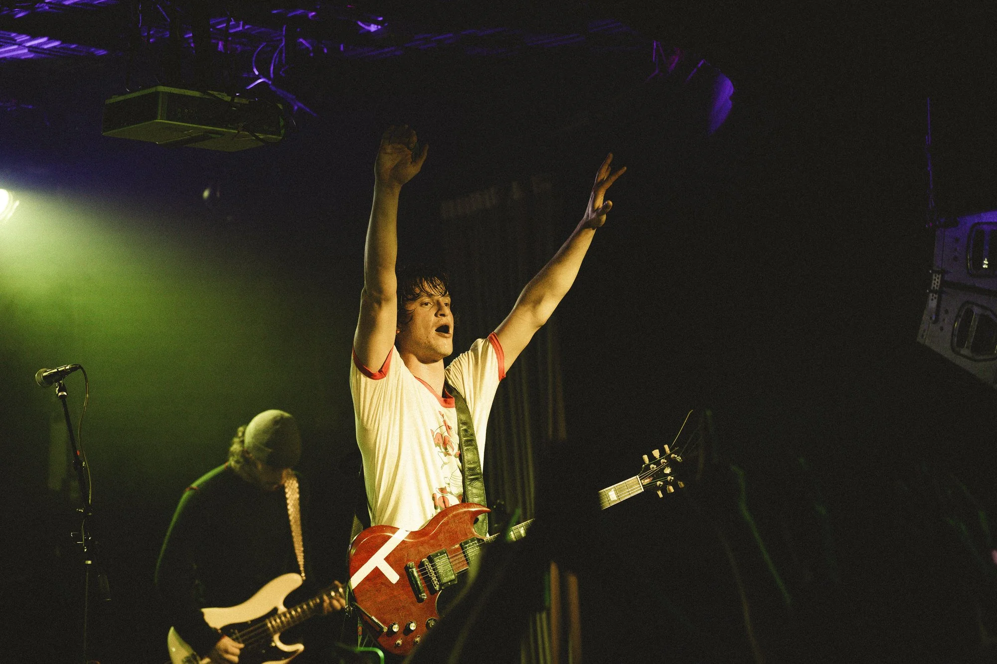 A male guitarist on stage with his arms raised, singing passionately. Another musician plays guitar in the background under green stage lighting.