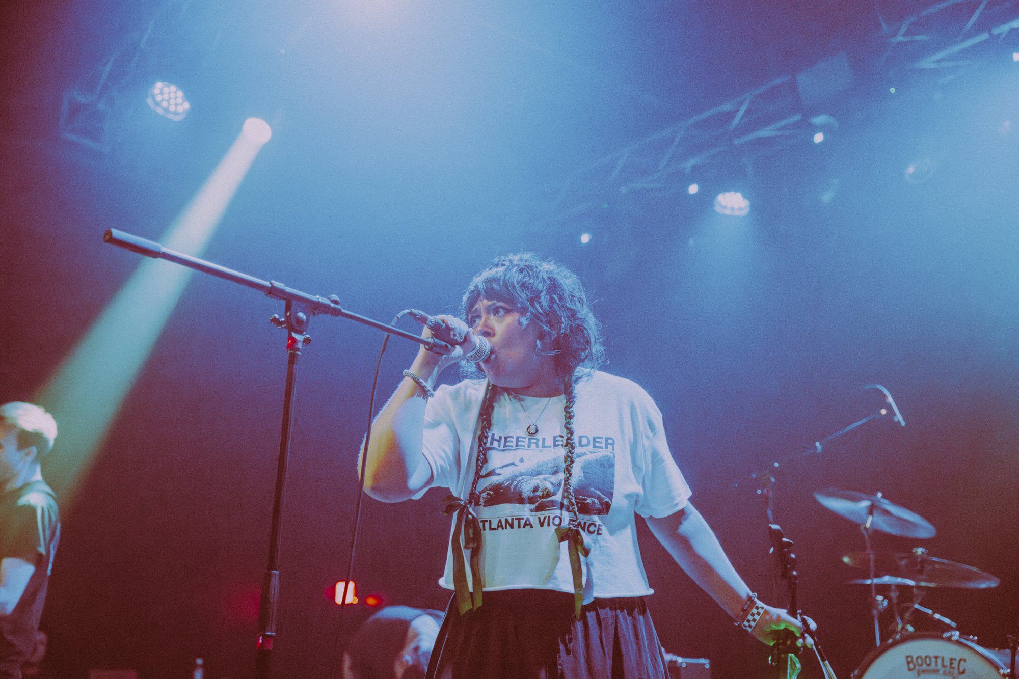 A young woman with black braided hair singing into a microphone on stage under blueish-purple lighting, wearing a white T-shirt with text, surrounded by other musicians and musical equipment.