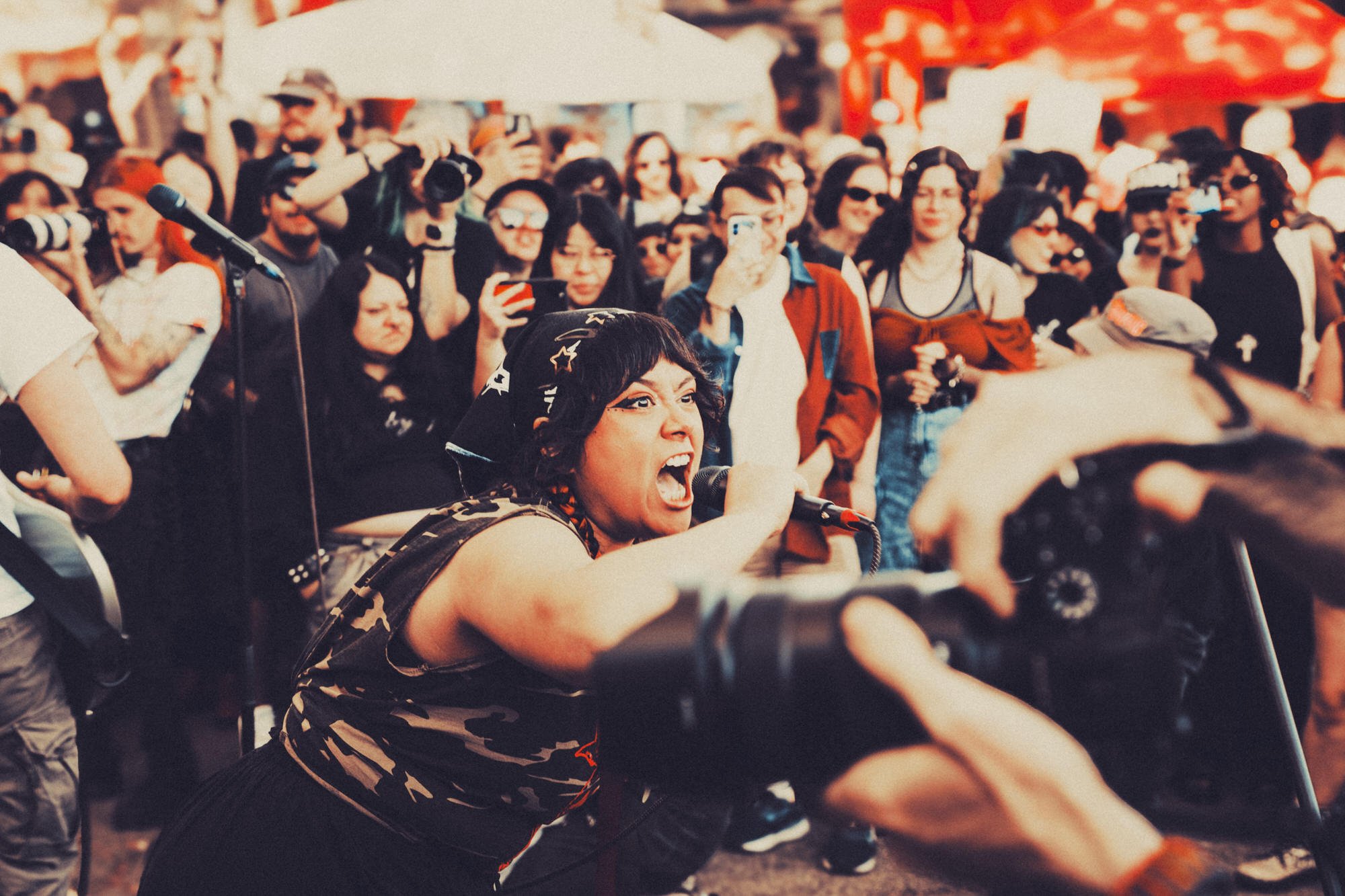A woman passionately singing into a microphone during a protest or rally, surrounded by a crowd of people with some holding cameras and signs.