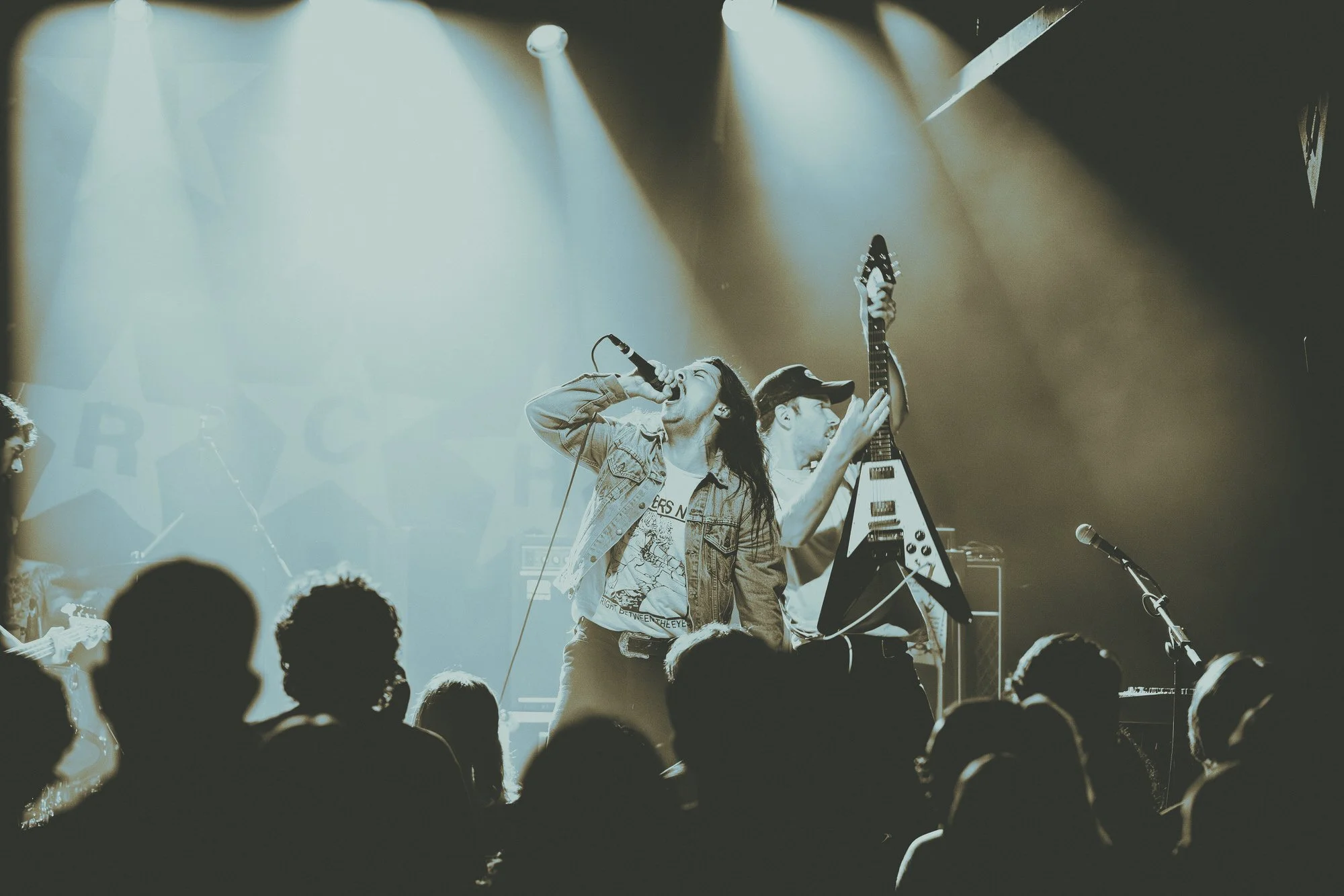 A band performs on stage with bright spotlights, featuring a singer with long hair singing into a microphone, and a guitarist holding an electric guitar. Audience silhouettes are visible in the foreground.