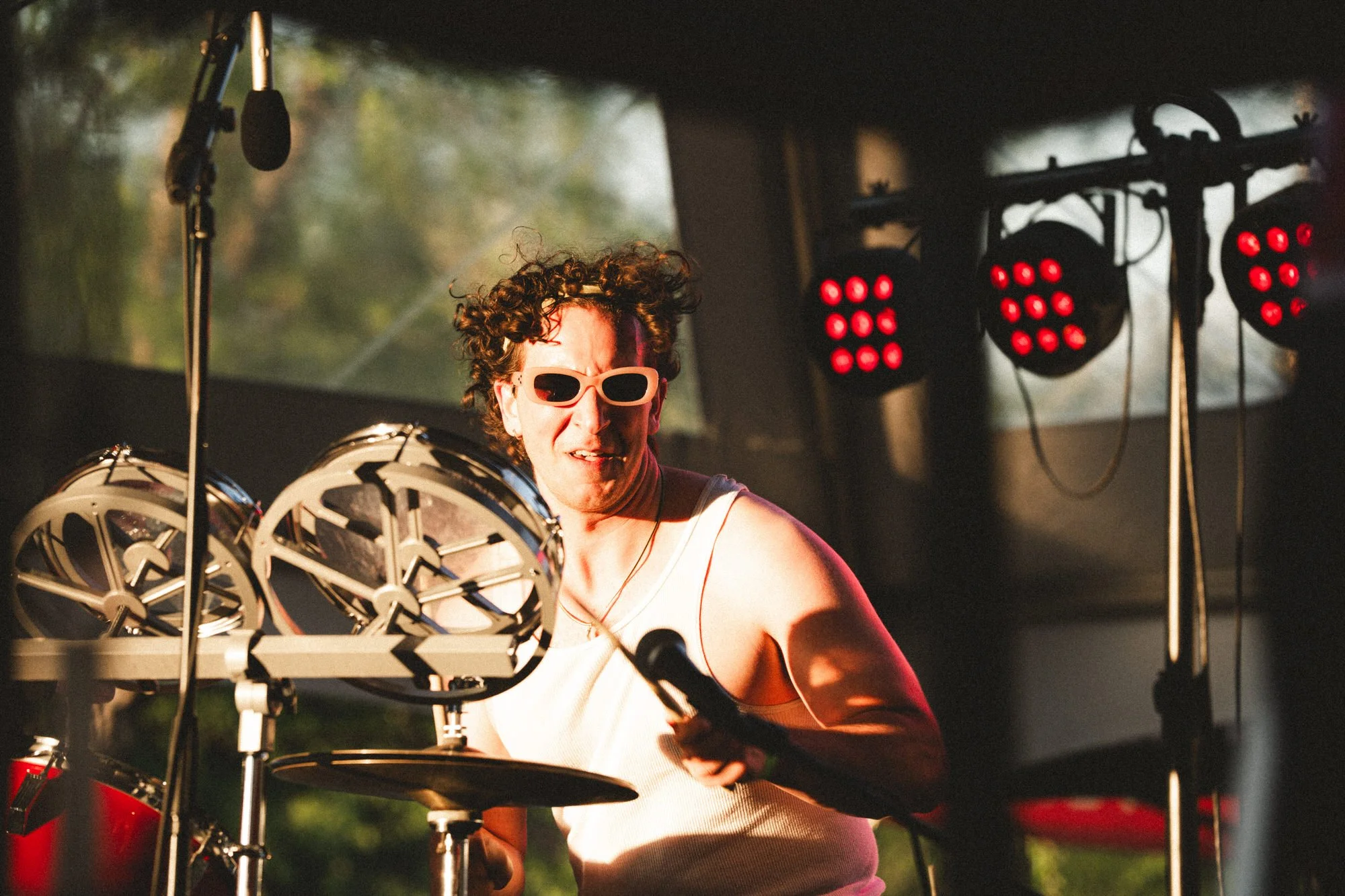 A person with curly hair wearing sunglasses, playing a drum set with a microphone and stage lights in the background.
