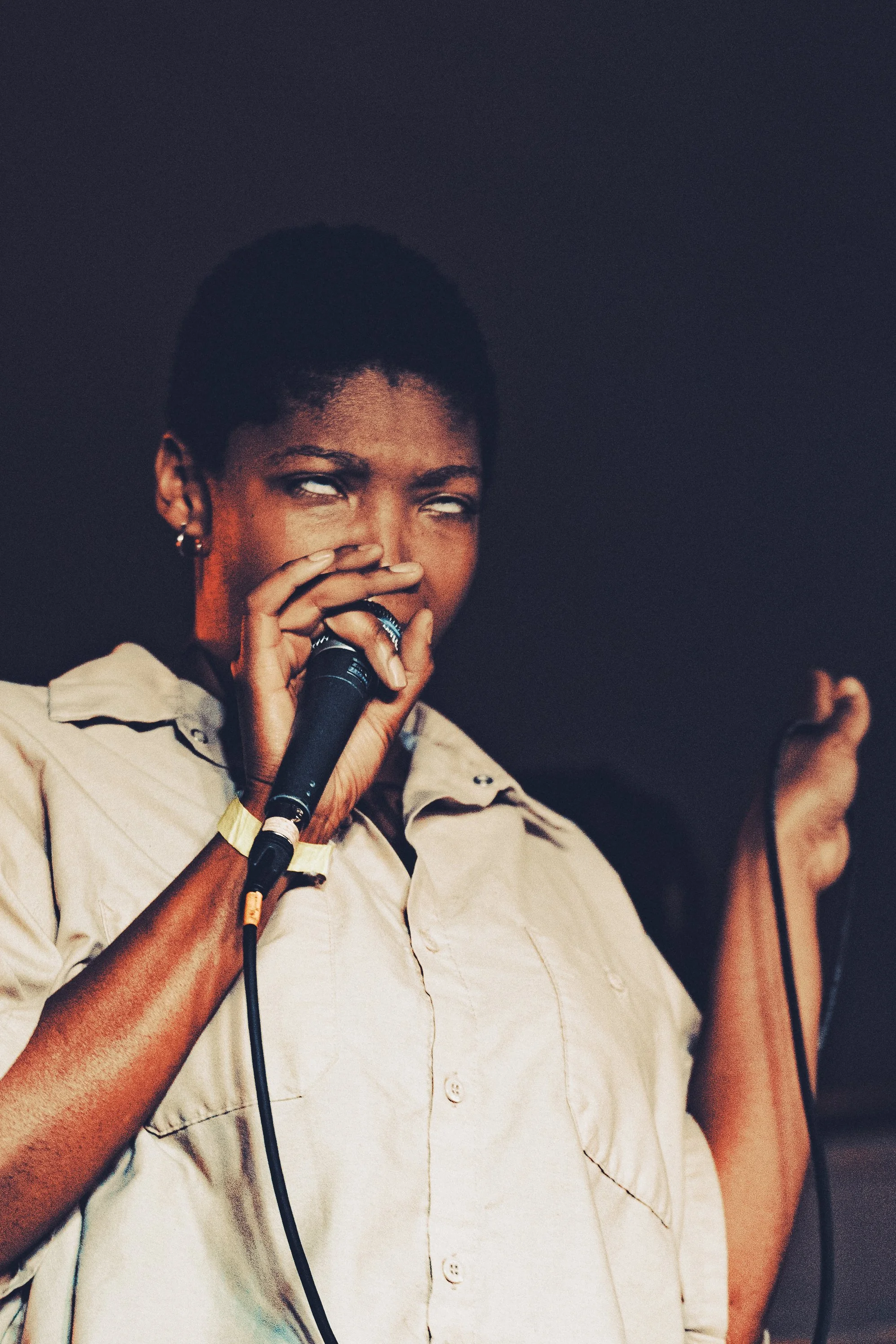 A woman with short hair singing into a microphone, wearing a beige shirt, with a dark background.