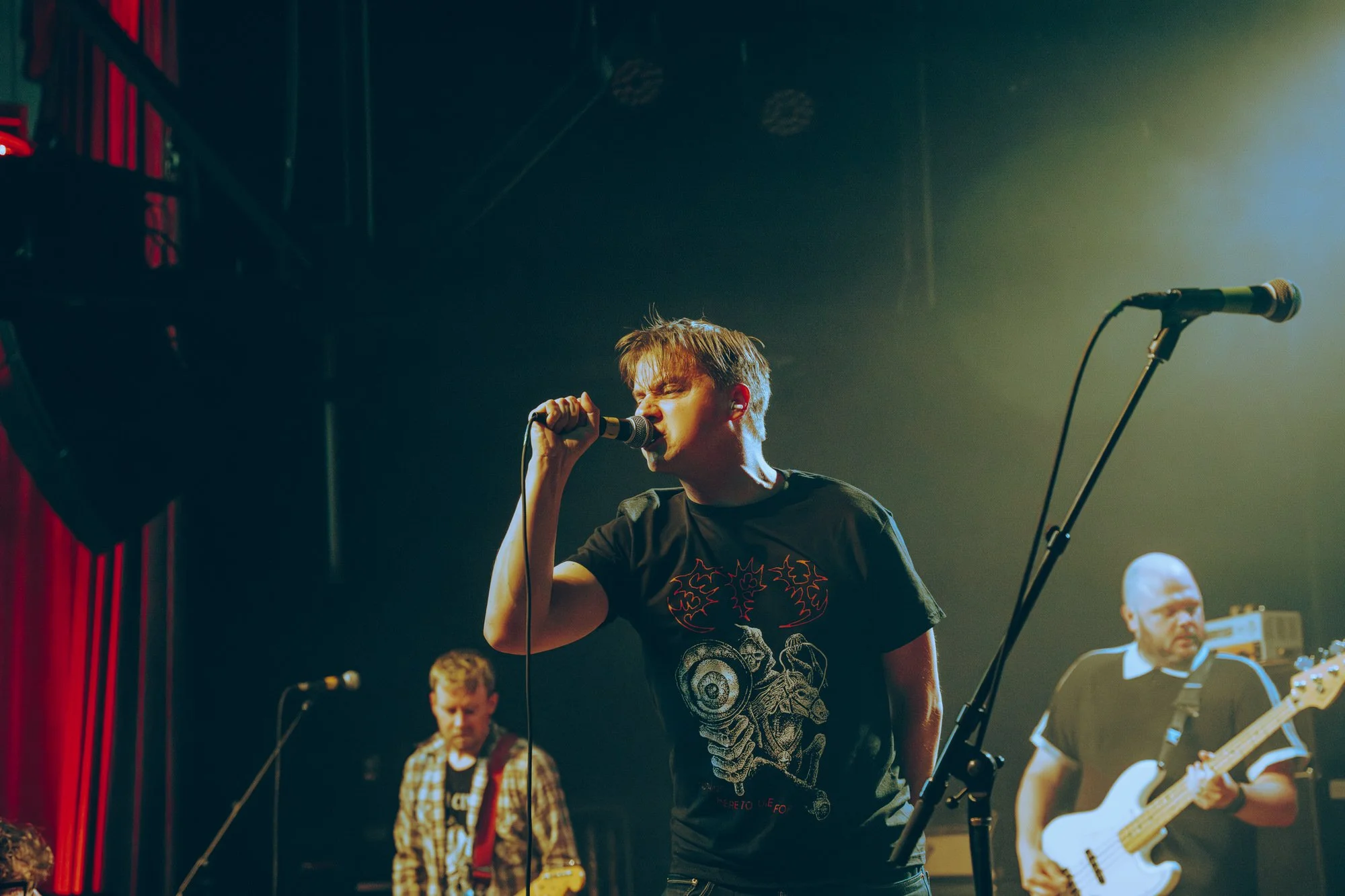 A young man passionately singing into a microphone on stage during a live band performance. Background musicians playing guitars and drums, with a dark stage lit by colorful stage lights.