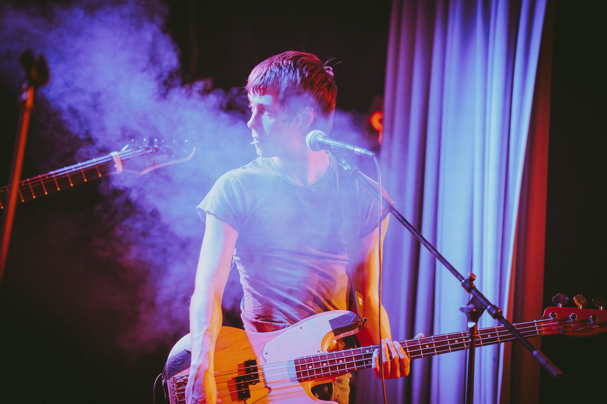 A young man with short hair playing a bass guitar on stage surrounded by purple smoke and blue stage lighting.