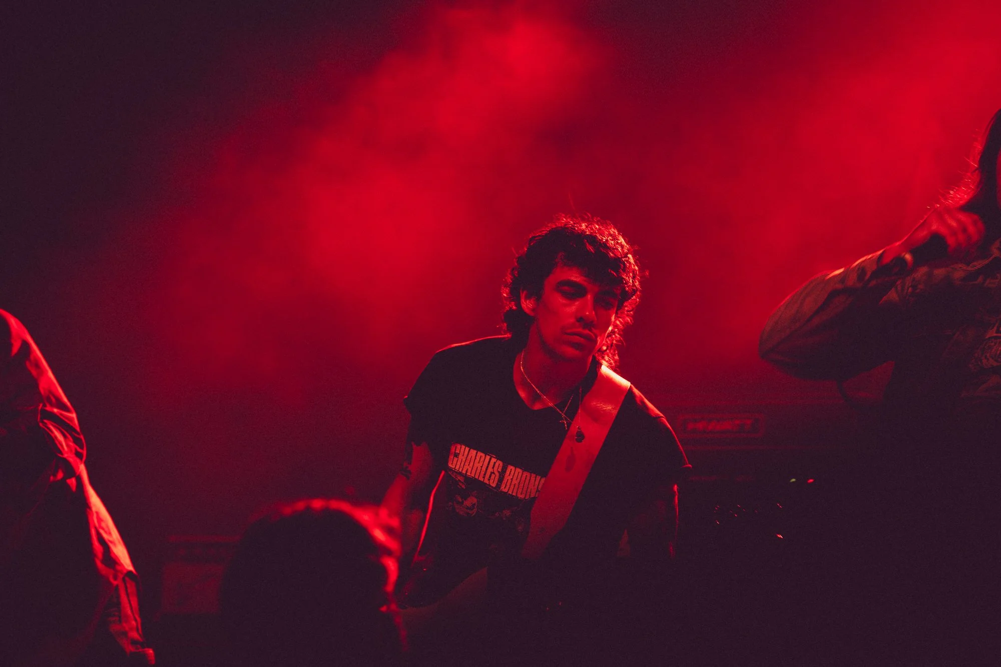 A person with curly hair and a black T-shirt with red and white text, standing in a red-lit environment.