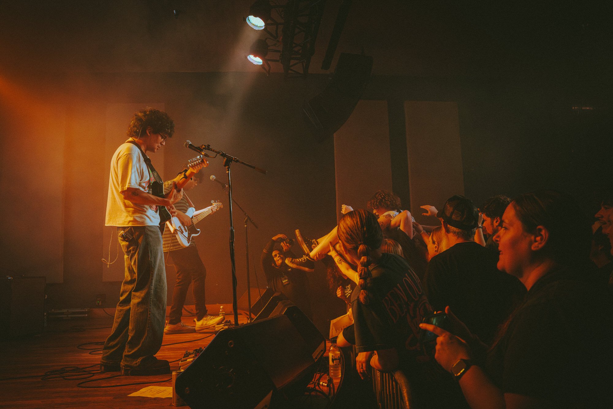 A band performs on stage with members playing guitars and singing, while the audience stands close, some reaching out or dancing.