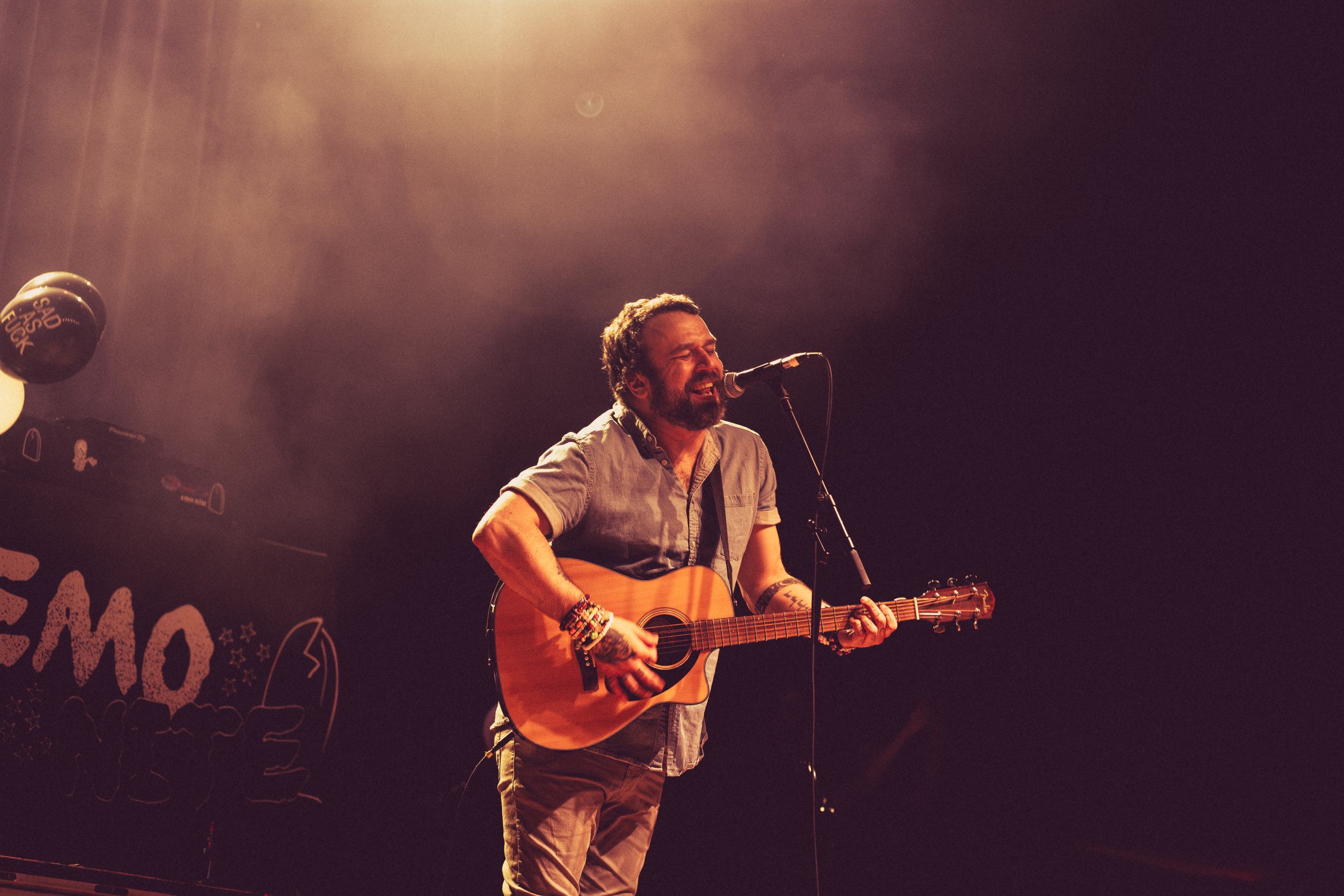 A man with a beard and tattoos singing into a microphone while playing an acoustic guitar on stage, smiling with closed eyes.
