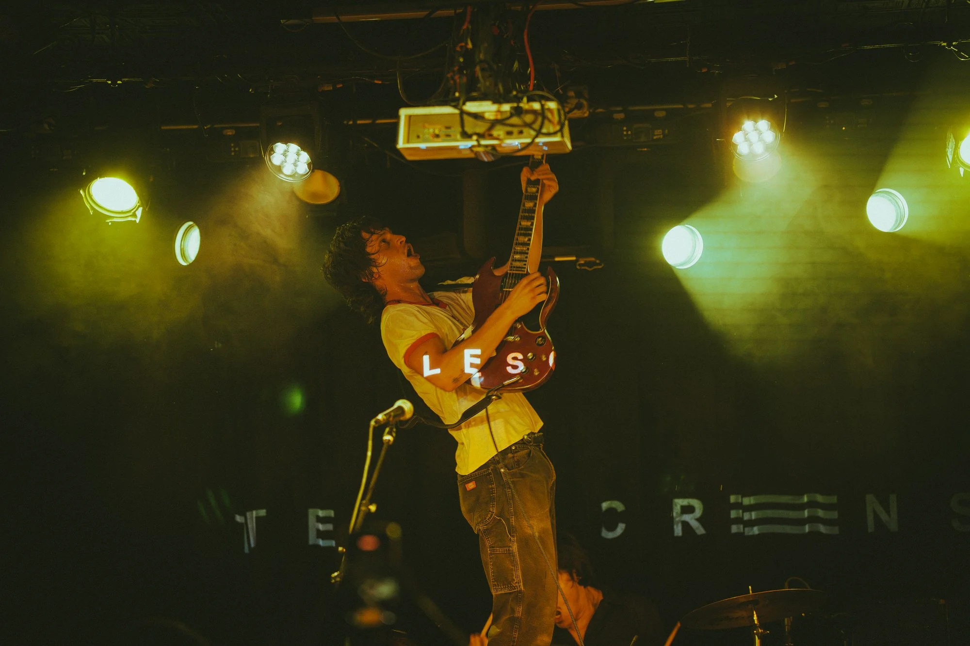 A musician on stage playing an electric guitar, illuminated by yellow and white stage lights, with a dark background and partially visible letters spelling 'TEEN CREEENS'.