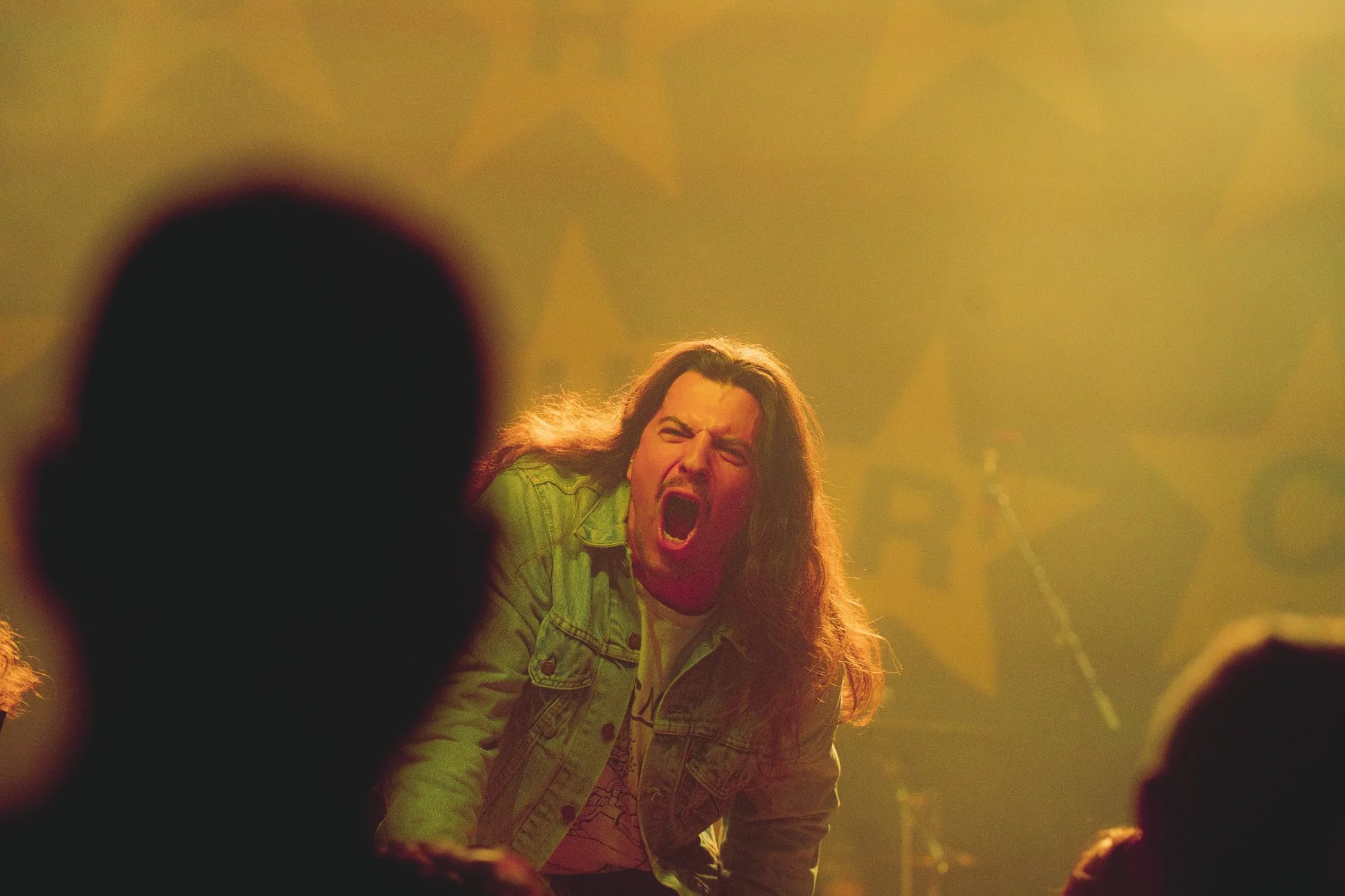 A woman with long hair and a green jacket is singing or shouting passionately on stage, surrounded by yellow-green lighting and blurred figures in the foreground.