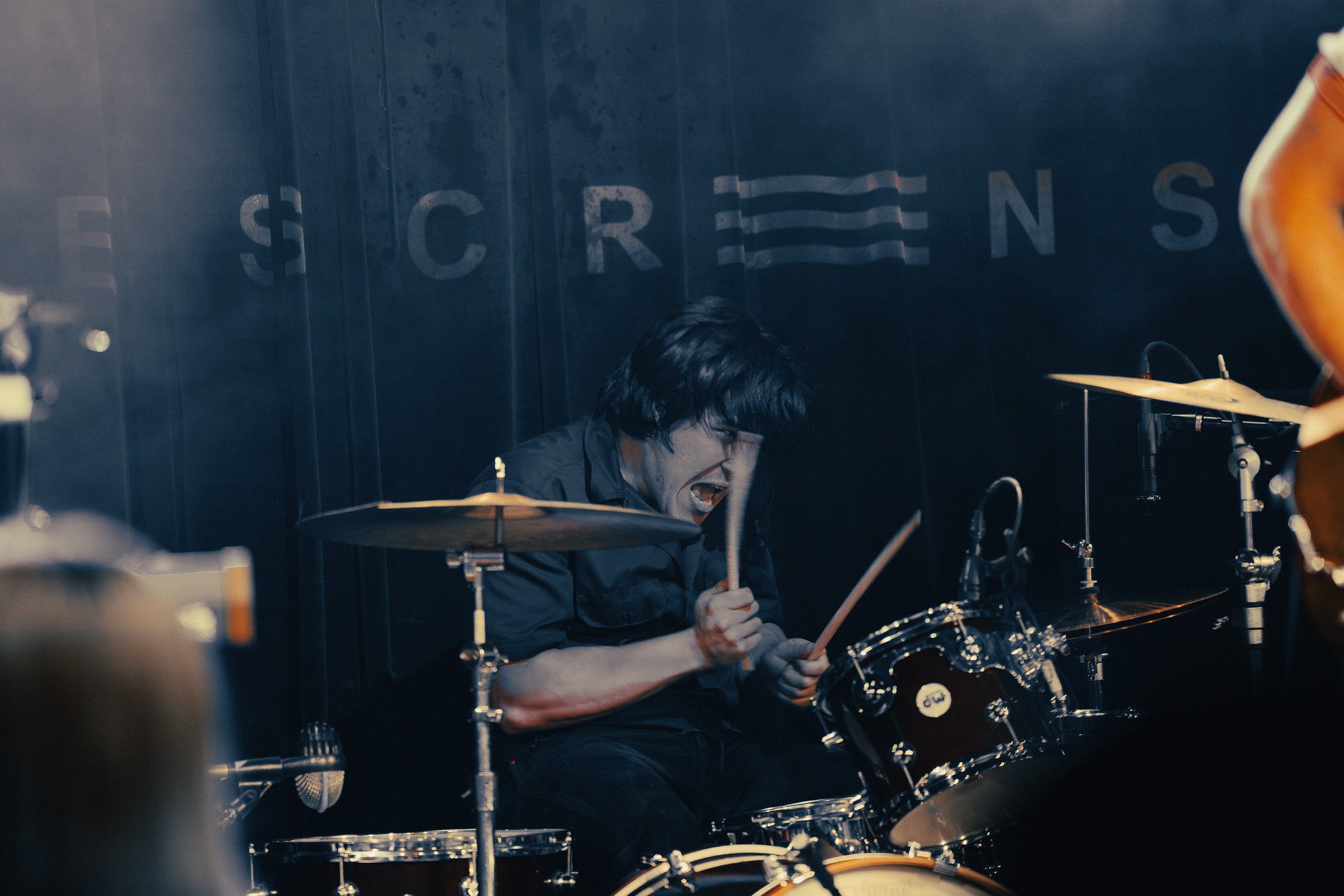 A drummer passionately playing a drum kit on stage, with dark background and part of a sign visible that spells out 'SCREEN'.